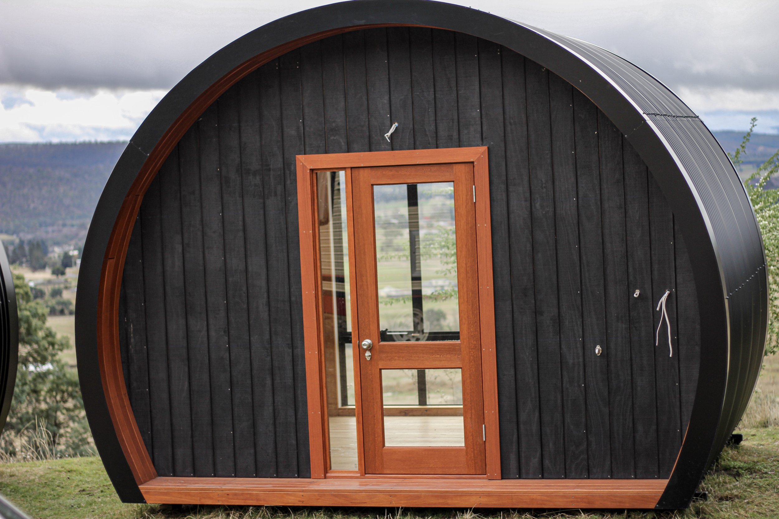A small, rounded wooden tiny house with black exterior siding, a wooden door with glass and a screen door, and a view of distant mountains and cloudy sky in the background.