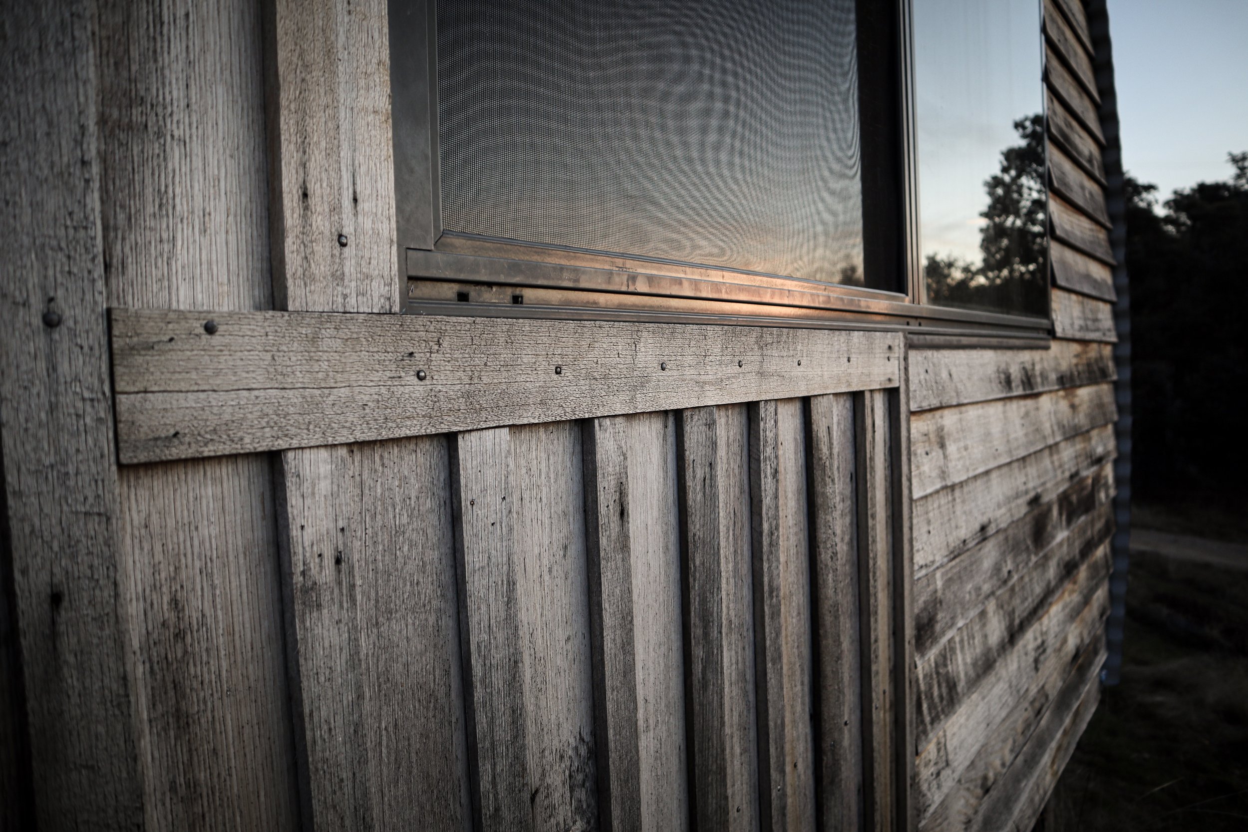 Close-up of weathered wooden house siding with a window reflecting trees and sky during sunset.