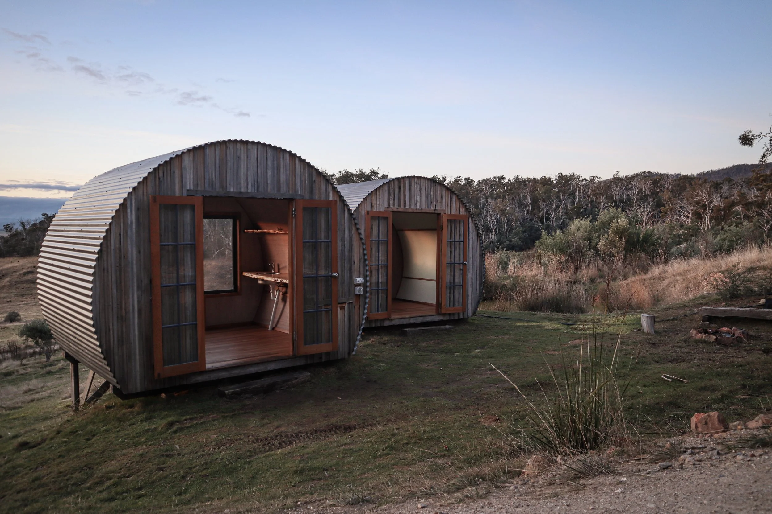 Two small, rounded wooden cabins with glass-paneled doors, situated on a grassy hillside with a natural landscape and trees in the background at dusk.