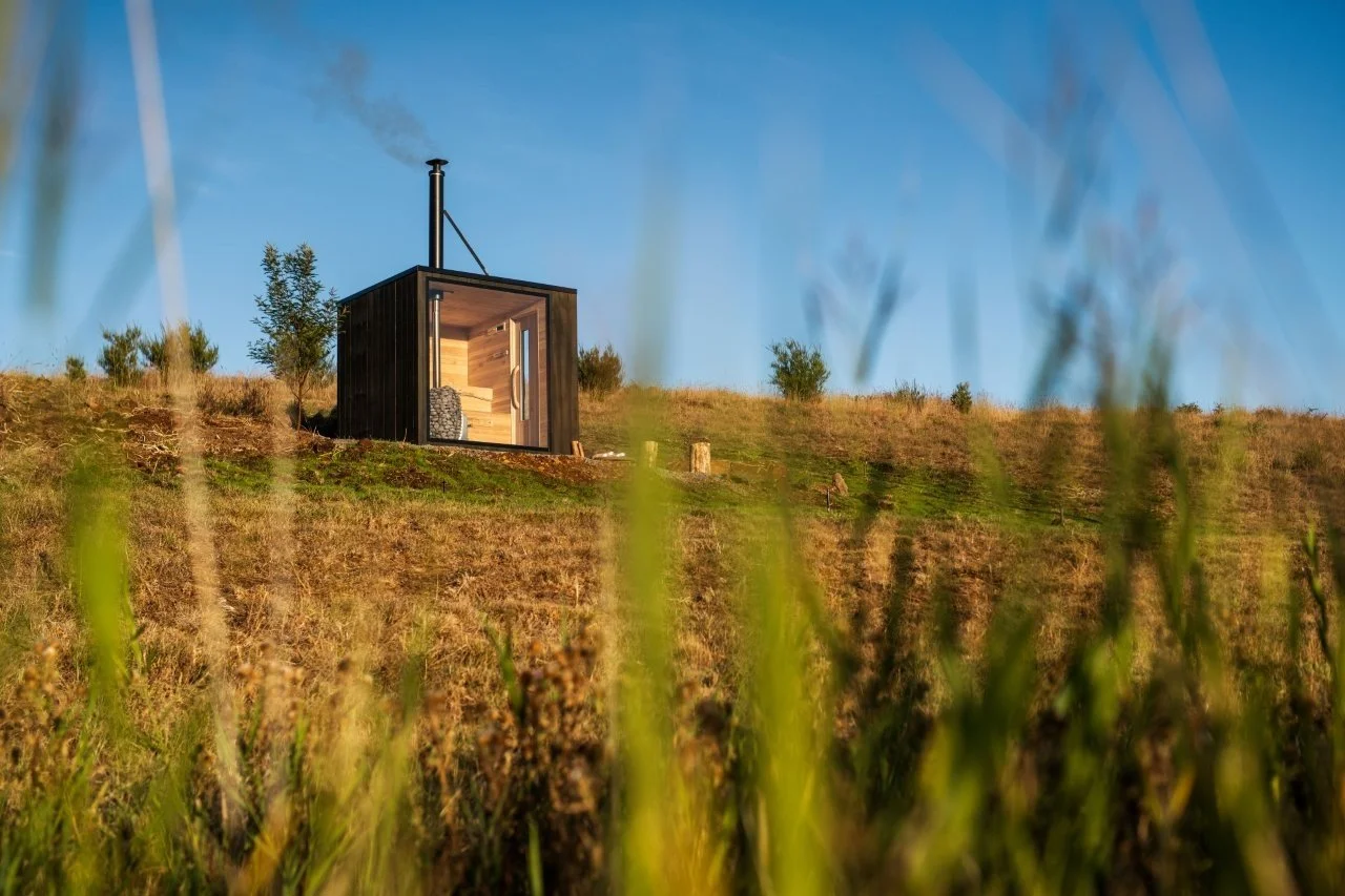 Small black cabin with a chimney on a grassy hill under a blue sky, with blurred grass in the foreground.