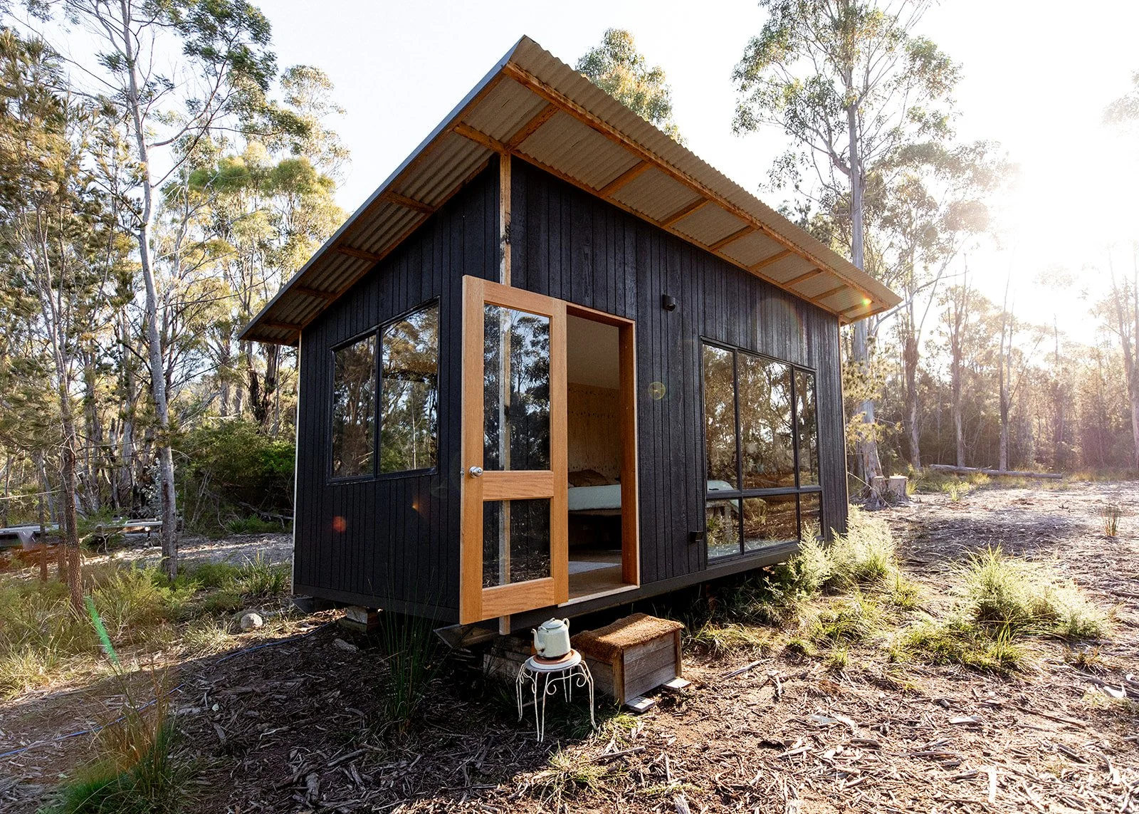 Small modern cabin with black wooden siding and large glass windows, situated in a wooded area with sunlight filtering through trees.