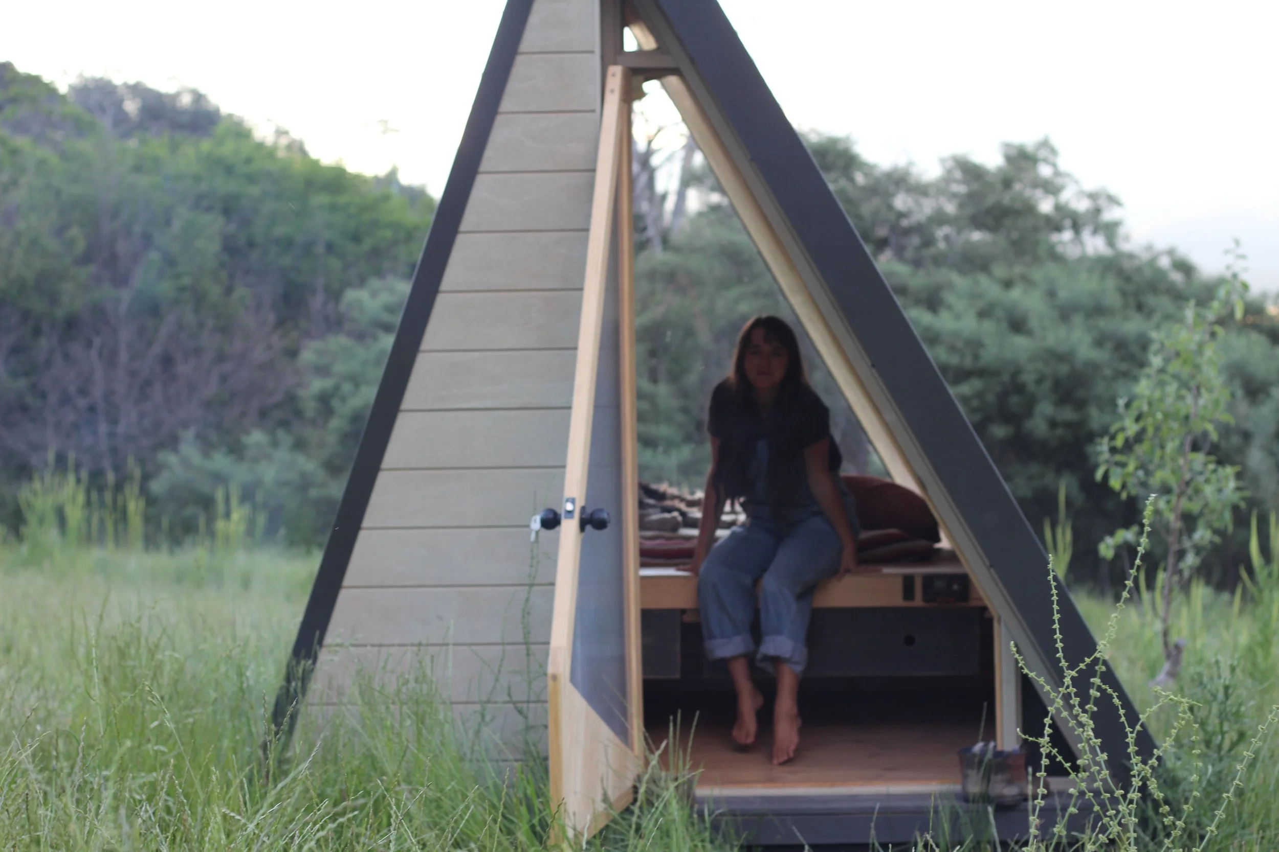A woman sitting on the bed inside an A-frame wooden tiny house, with a door open to a grassy outdoor area and trees in the background.