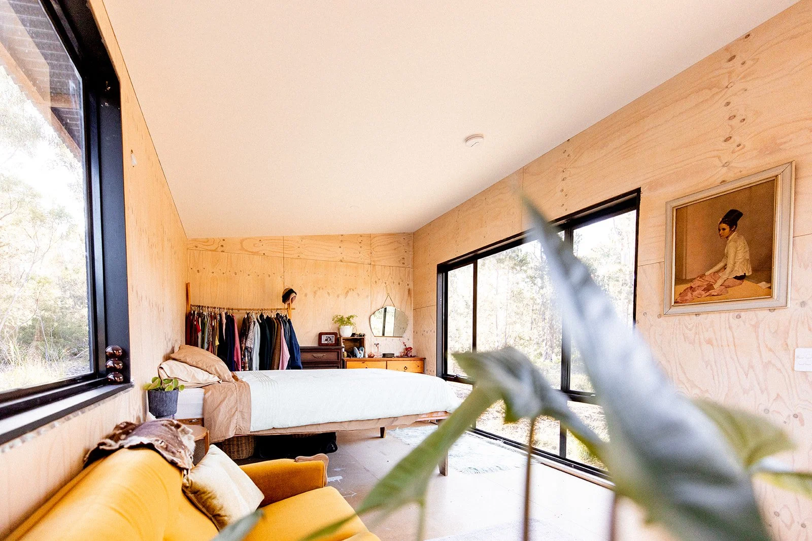 Room with large windows, bedside table, bed, clothes hanging on a rack, dresser, mirror, painting of a woman, and a yellow sofa