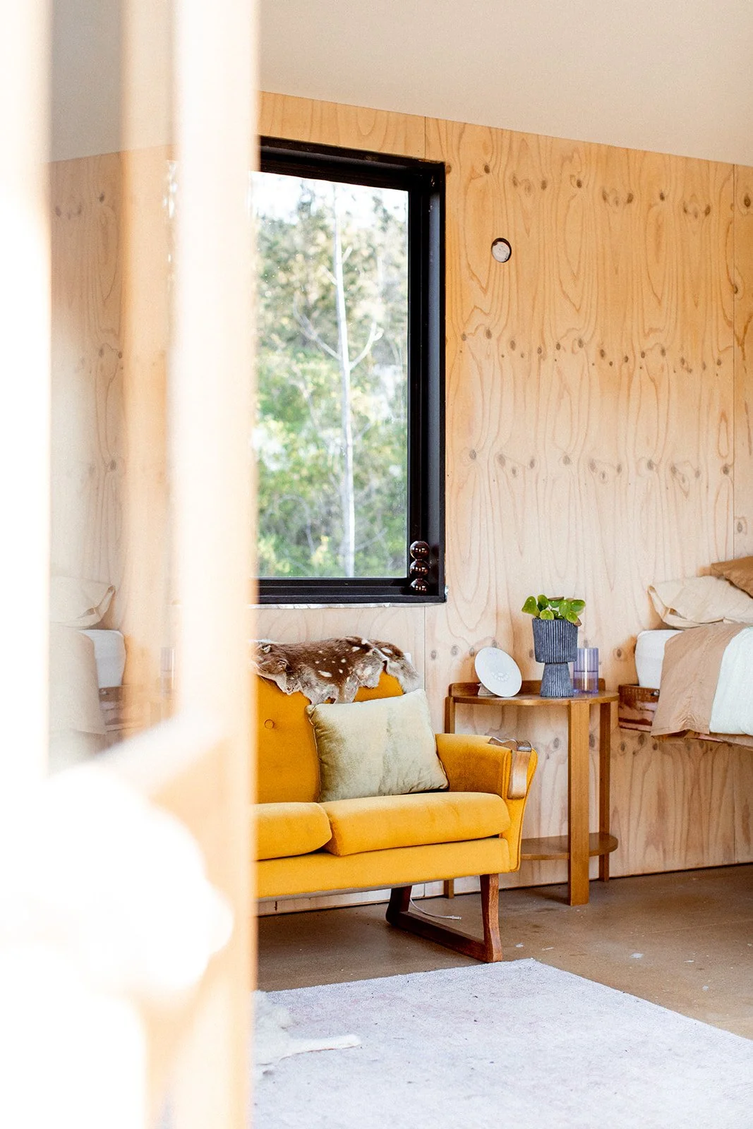 Interior of a room with natural wood paneled walls, a window showing greenery outside, a yellow sofa with throw pillows, a small wooden side table with a potted plant, a modern clock, and a partially visible elevated bed or platform with bedding.