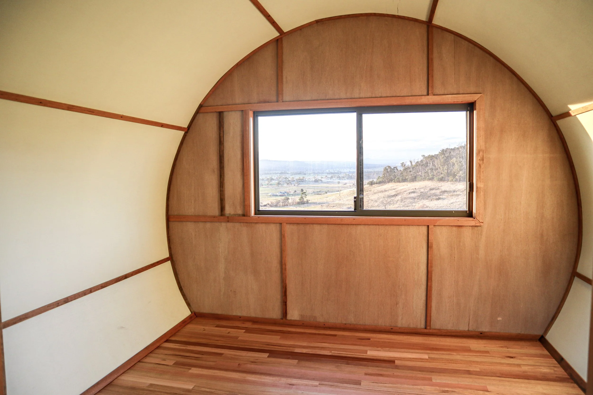 Interior of a small wooden room with a large window showing a landscape view of hills and fields.