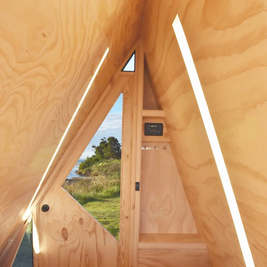 Interior of a triangular-shaped wooden tiny house or cabin with LED lighting, a door with a window, and a scenic outdoor view.