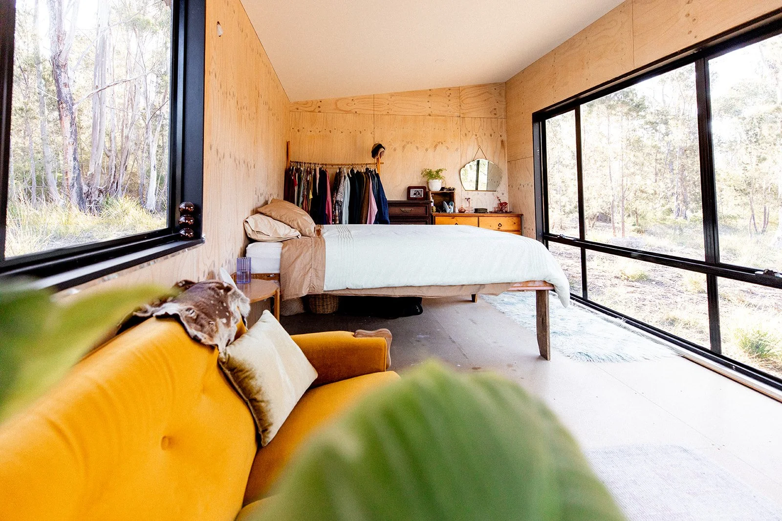 A bedroom with a large window overlooking a wooded landscape, a bed with beige bedding, a wooden dresser, an open closet with clothes, and a yellow sofa with pillows and a blanket.