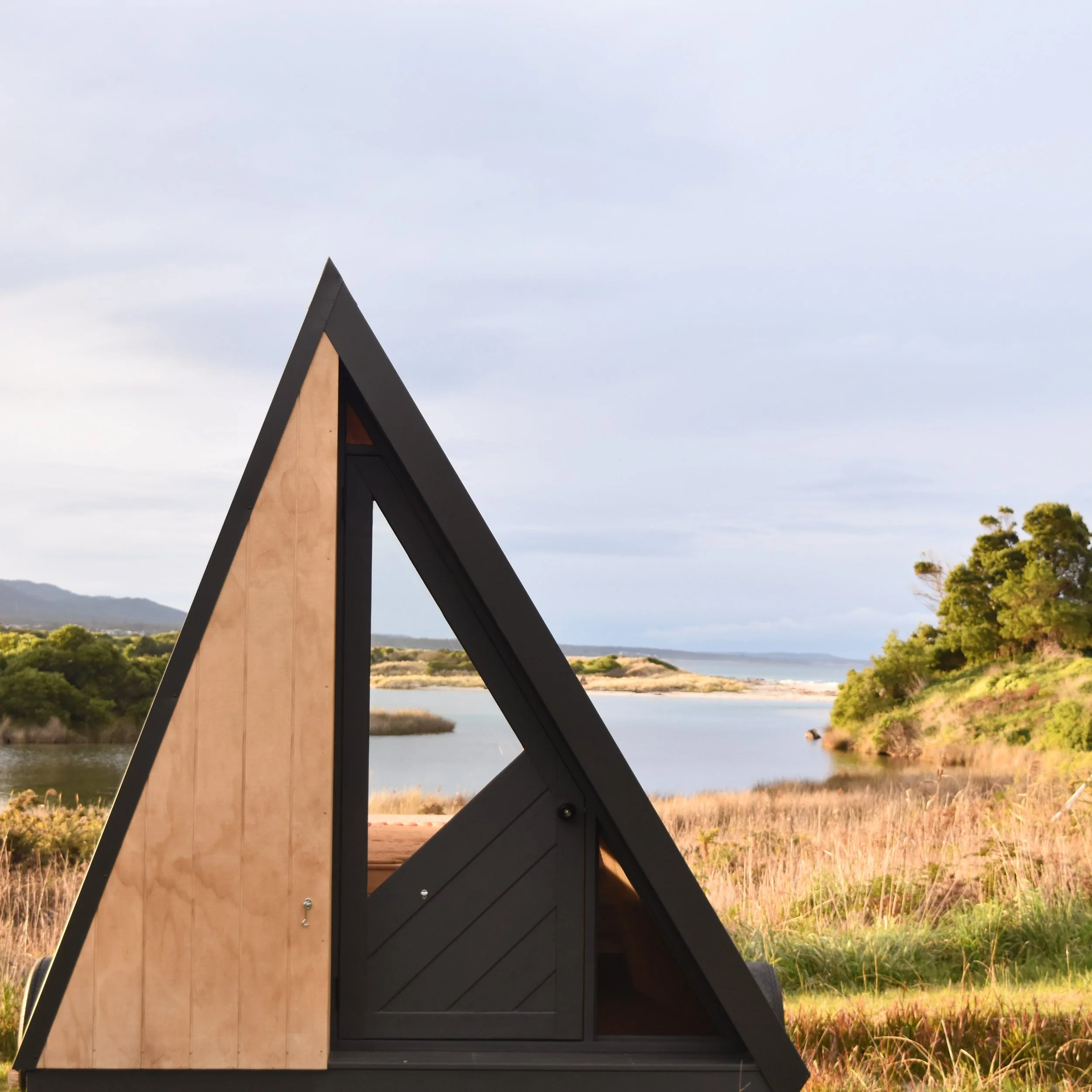 A small triangular wooden and black structure with a door, set outdoors near a lake with grassy areas, trees, and hills in the background under a cloudy sky.