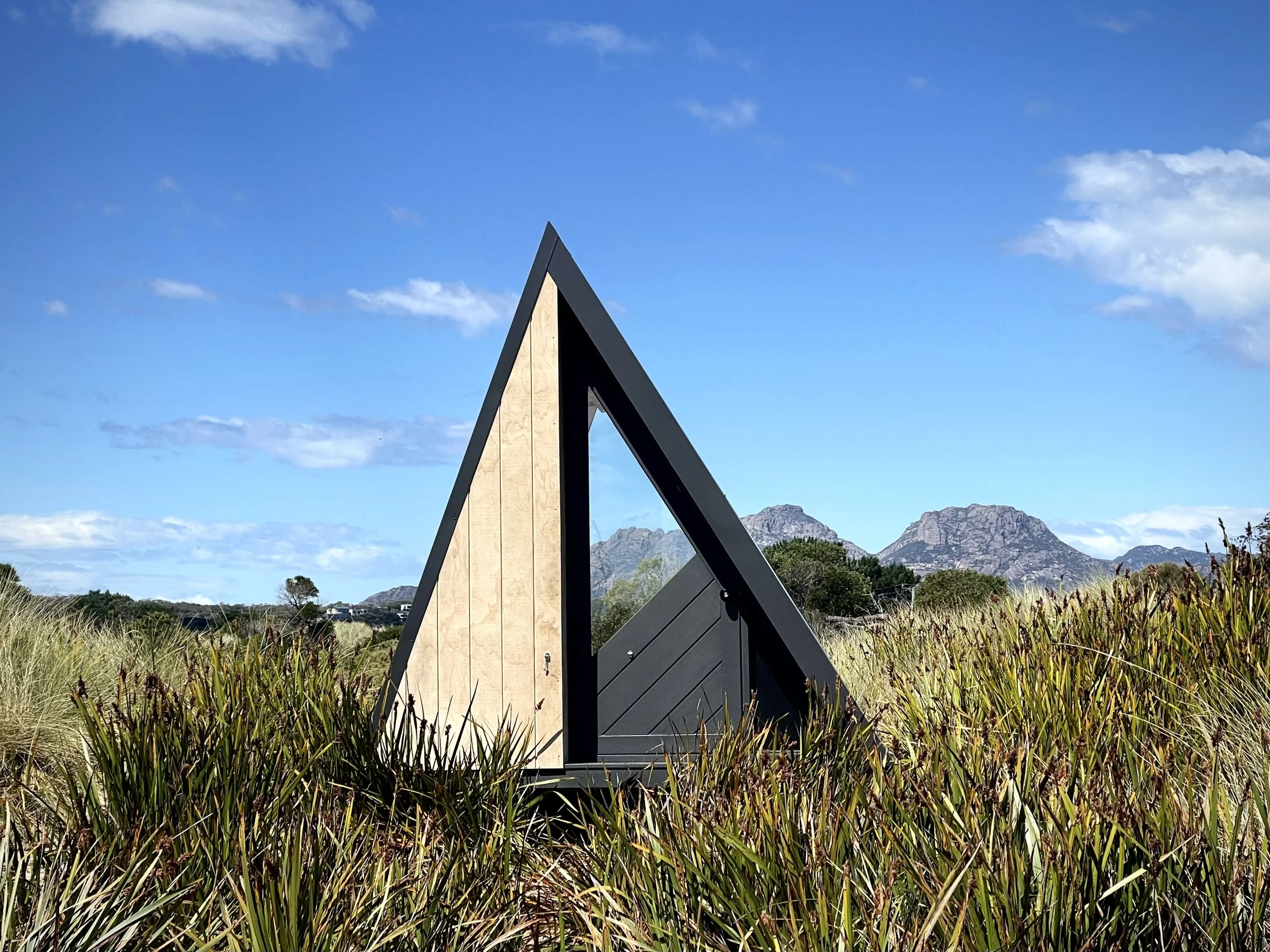 A triangular A-frame structure with wooden and black panel sections in a grassy field with mountains and a blue sky in the background.