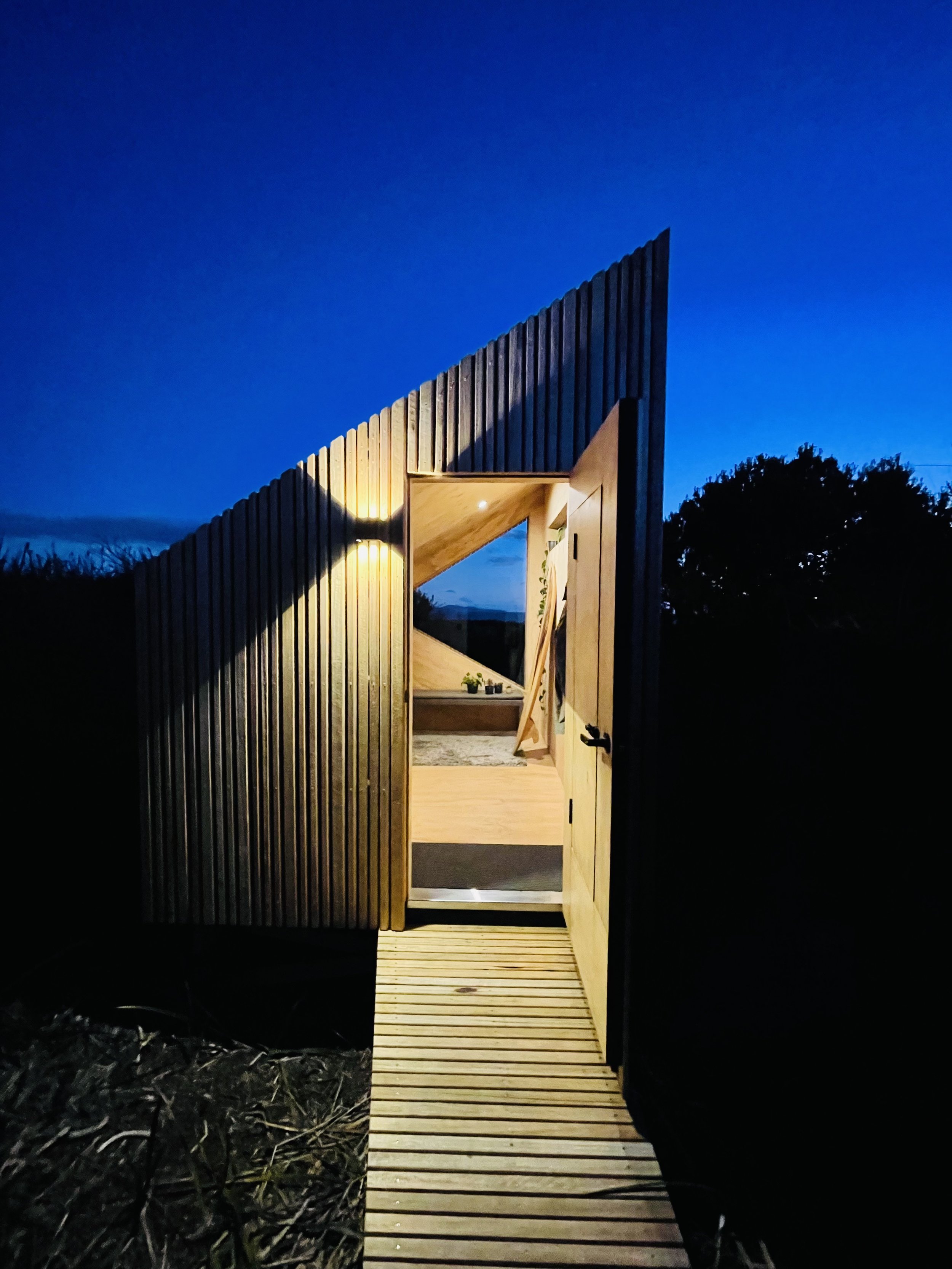 An exterior view of a small modern wooden cabin with vertical wooden siding, illuminated from inside, at dusk or nighttime. The entrance has a small wooden deck leading to a door, with a window on the front wall showing furniture and decor inside.