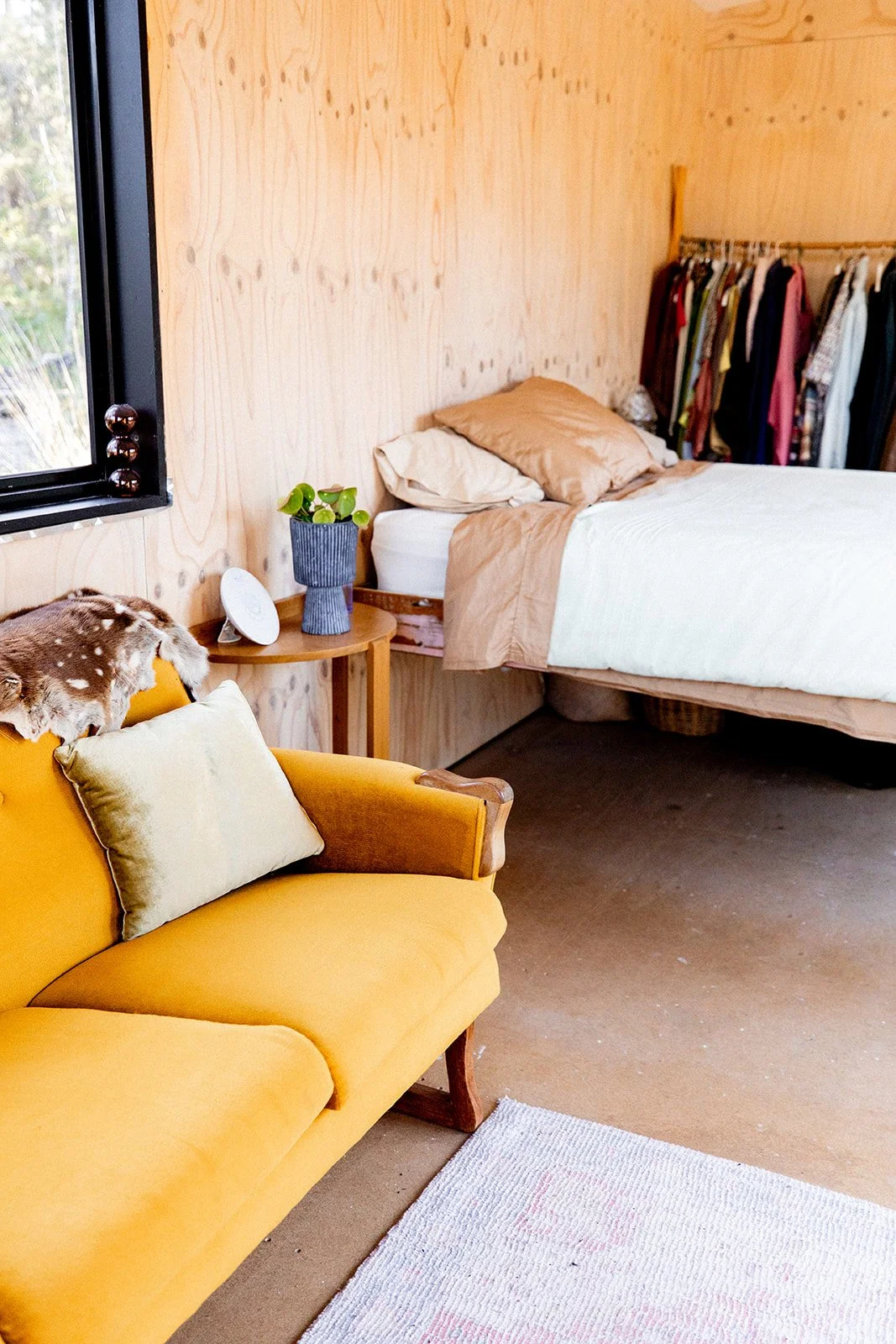 Interior of a bedroom with a mustard yellow armchair, a wooden side table with a potted plant, a bed with beige pillows, and an open closet with hanging clothes, next to a window.