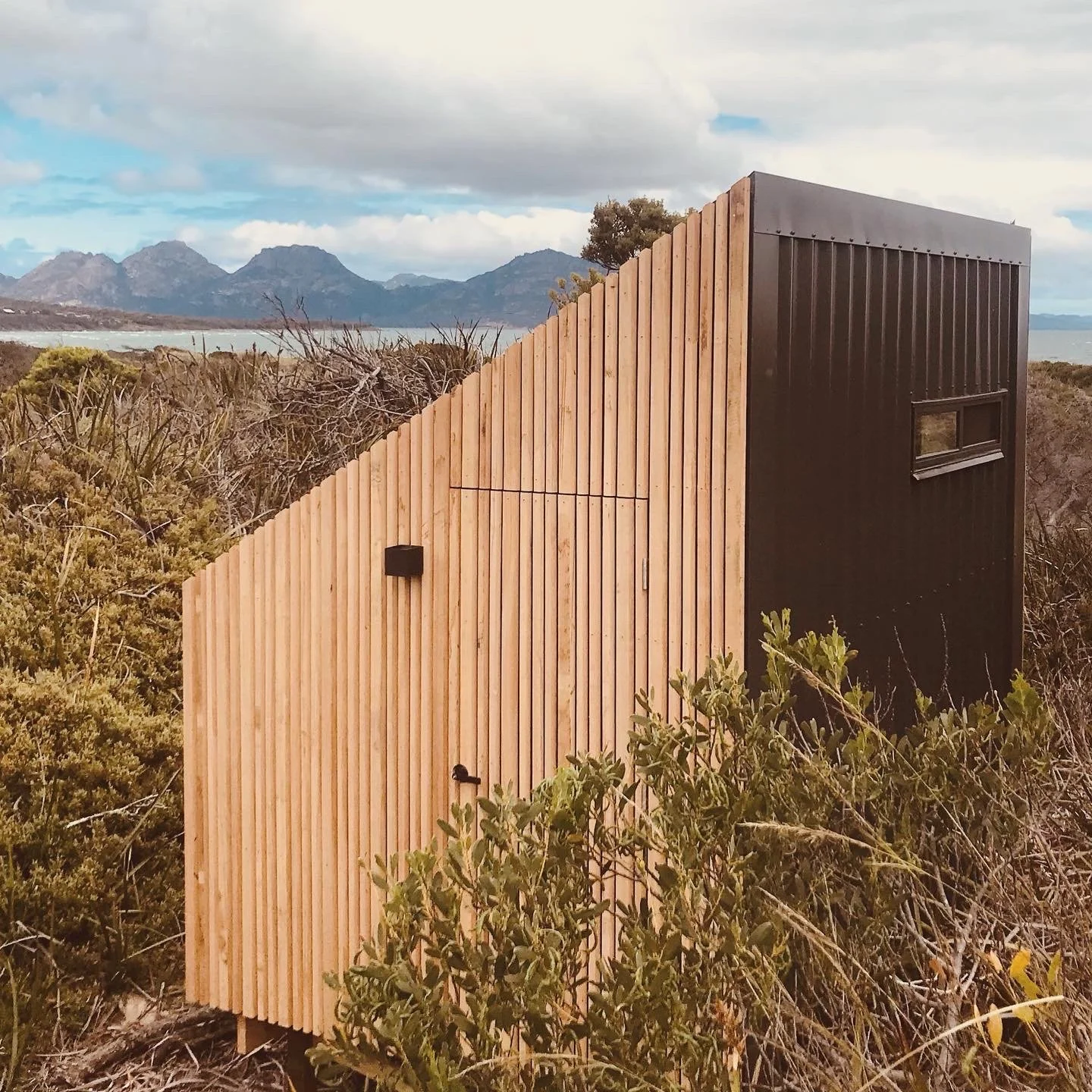 A modern small building with a sloped wooden facade on one side and black vertical siding on the other, located outdoors in a natural landscape with shrubs, mountains, and water in the background.