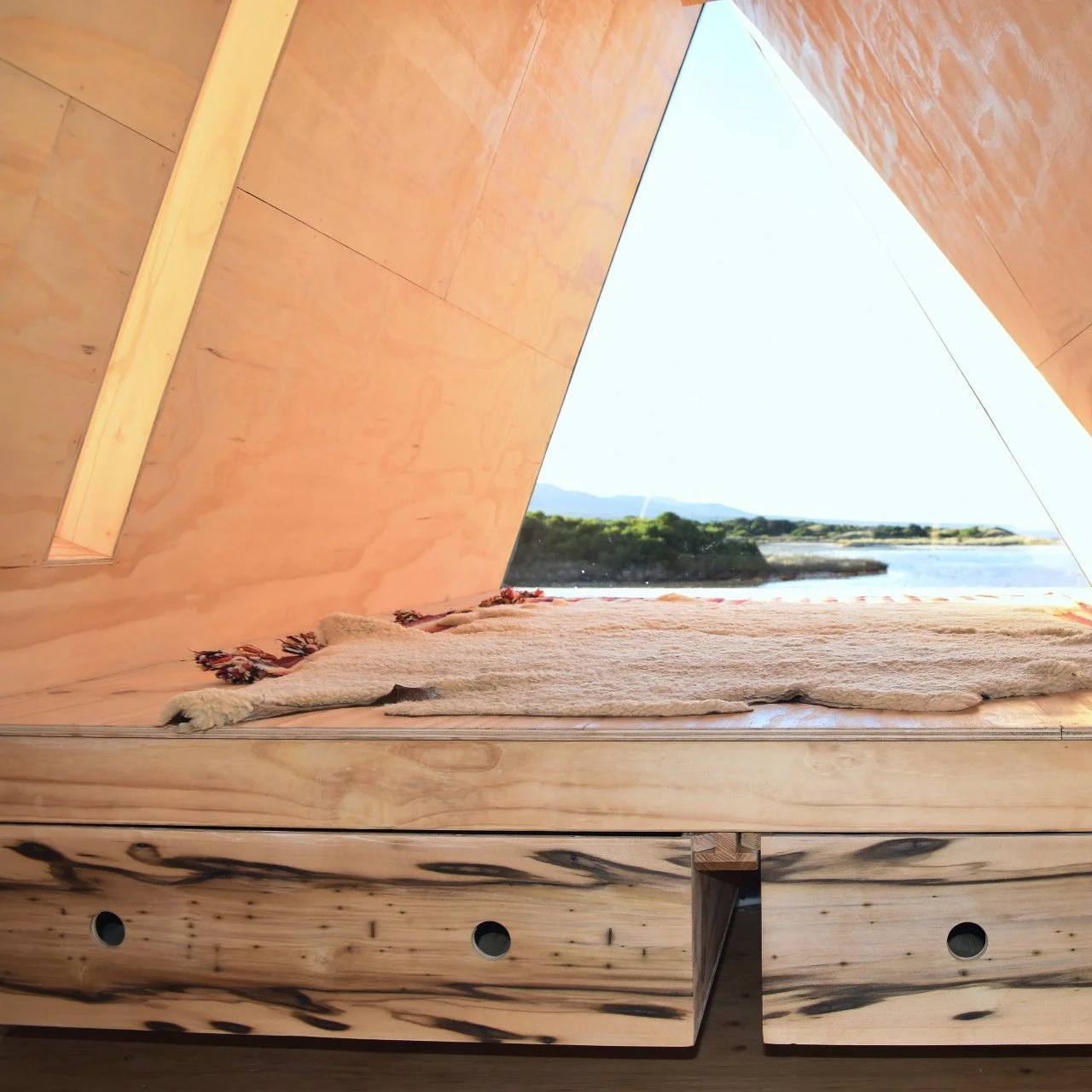 Inside of a room with angled wooden walls and a large triangular opening showing an outdoor landscape with water, trees, and distant mountains.