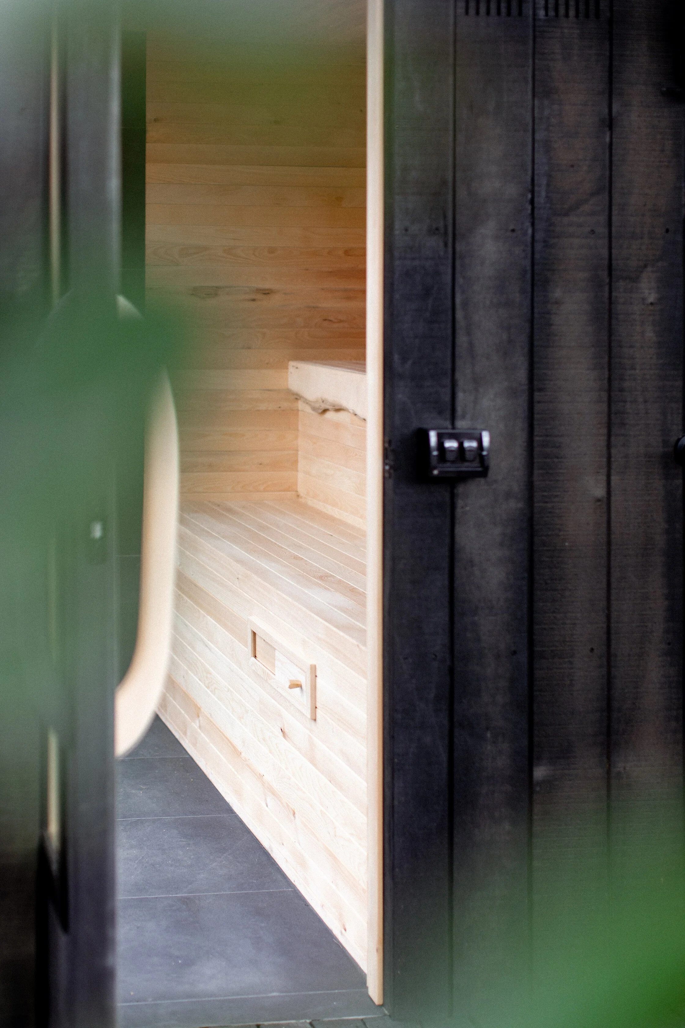 View into a wooden sauna room with light-colored wood paneling and a bench, partially obscured by a dark door with a lock.