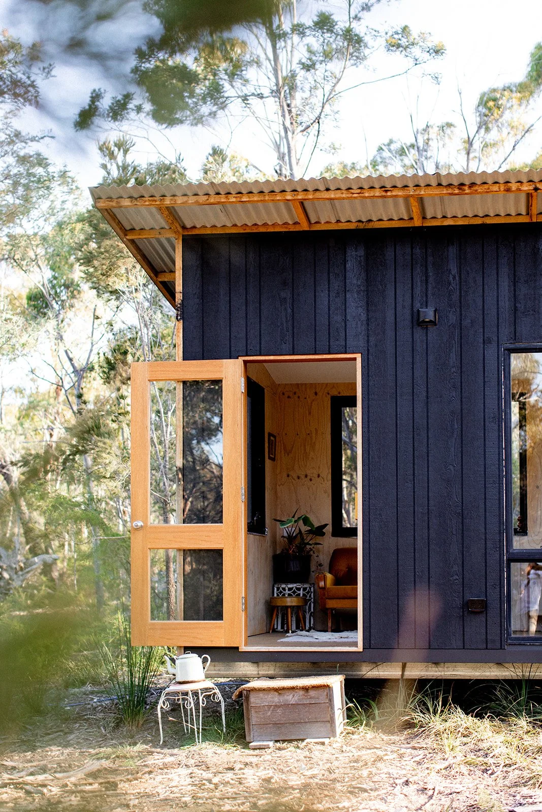 Small black wooden house with open wooden door and interior visible, surrounded by trees and outdoor plants, with a small white teapot on a decorative stool outside.