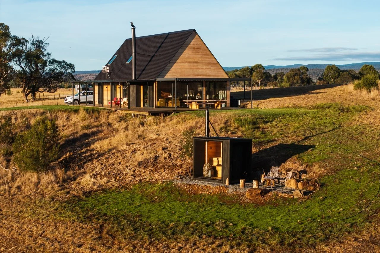 A modern house with a dark roof and large glass windows situated on a hill, with an outdoor seating area and a small black container-like structure in the yard, set in a rural landscape during daylight.