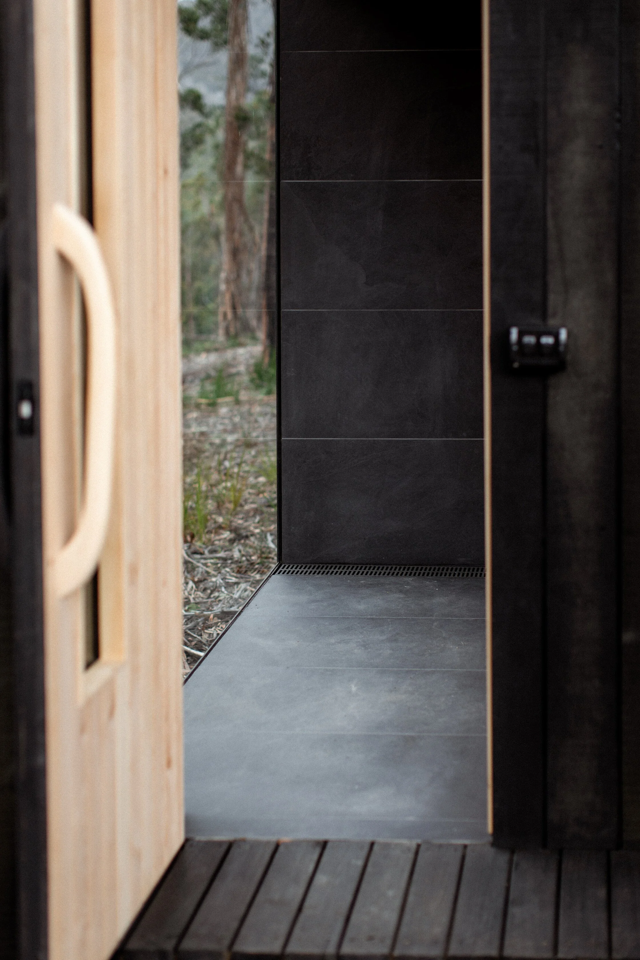 Close-up of black tiled walls and floor with a view outside through an open door showing trees and outdoor natural landscape.