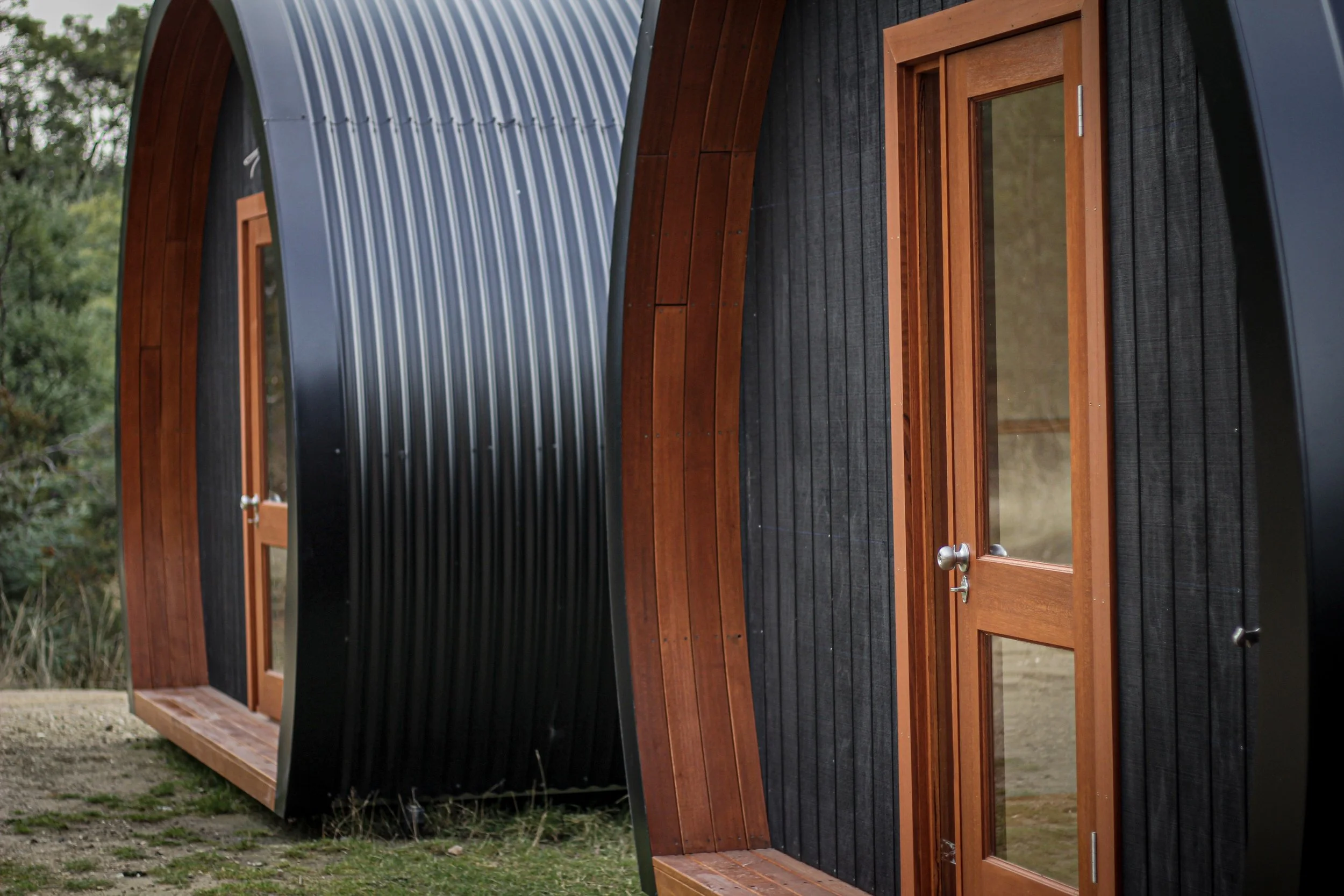 A small, modern, rounded tiny house with black metal siding and wood accents, featuring a glass door and a window, surrounded by greenery.