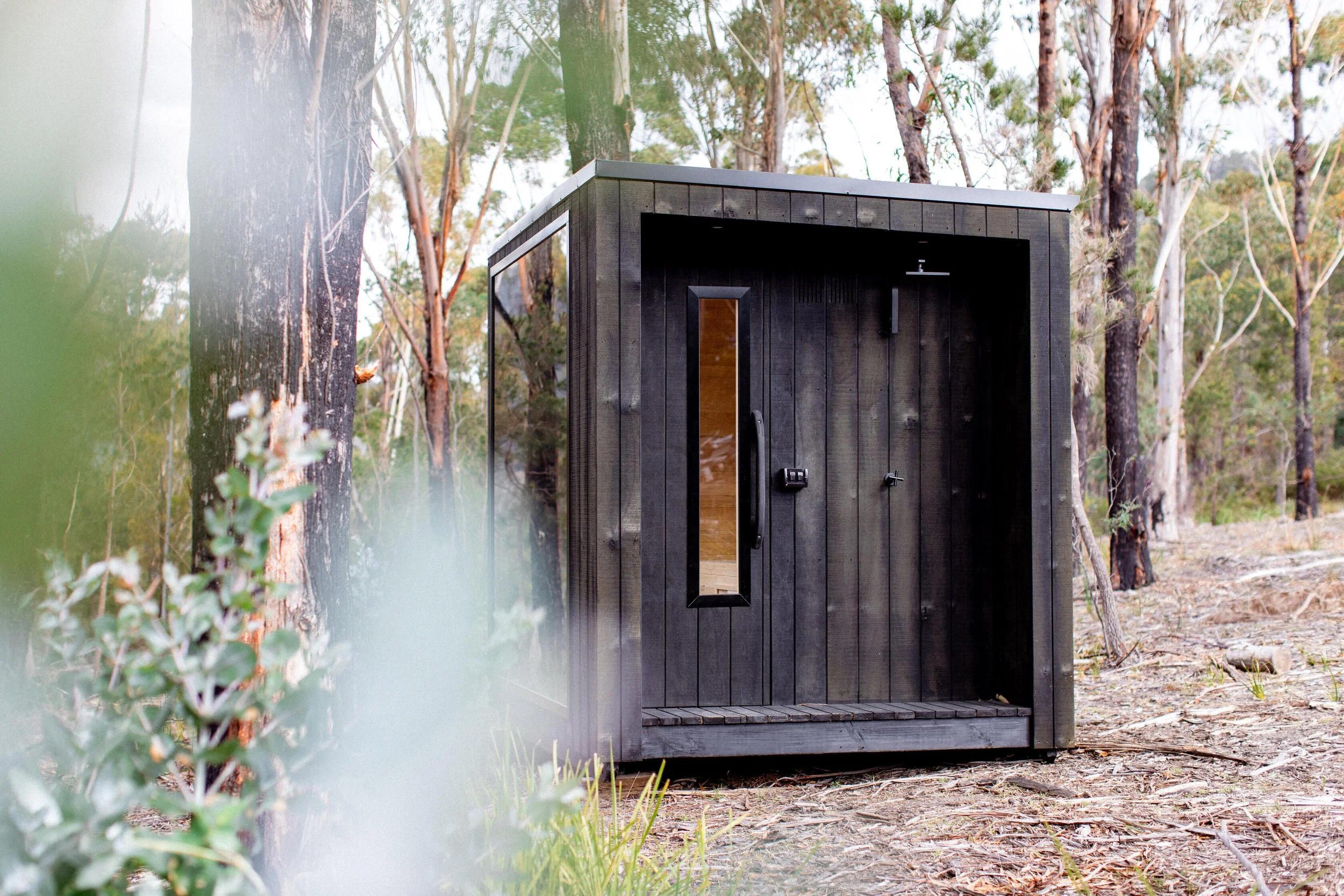 A black outdoor shower enclosure with a glass door is situated among trees in a forested area.