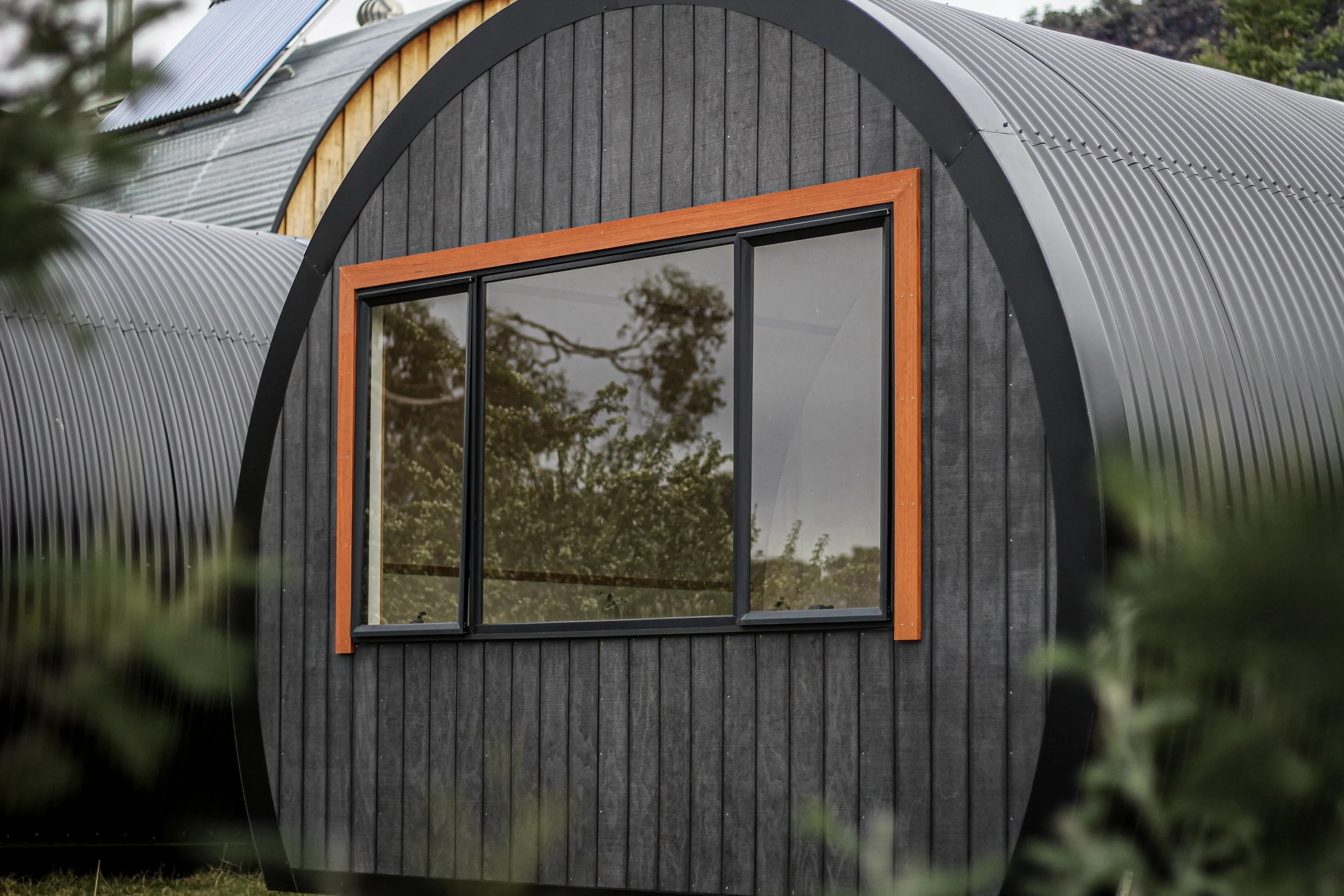 A modern tiny home with a black curved metal exterior, large sliding glass window framed with wood, and surrounded by greenery.