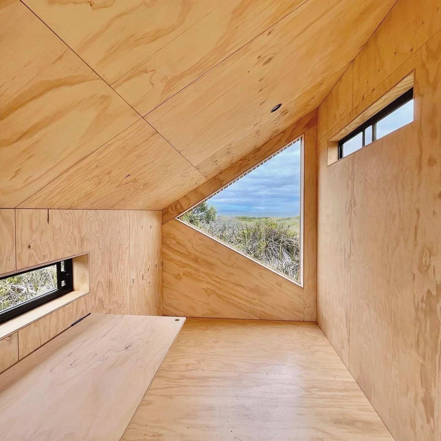 Interior of a small wooden room with angled ceilings and three narrow windows showing an outdoor landscape with grasses and cloudy sky.