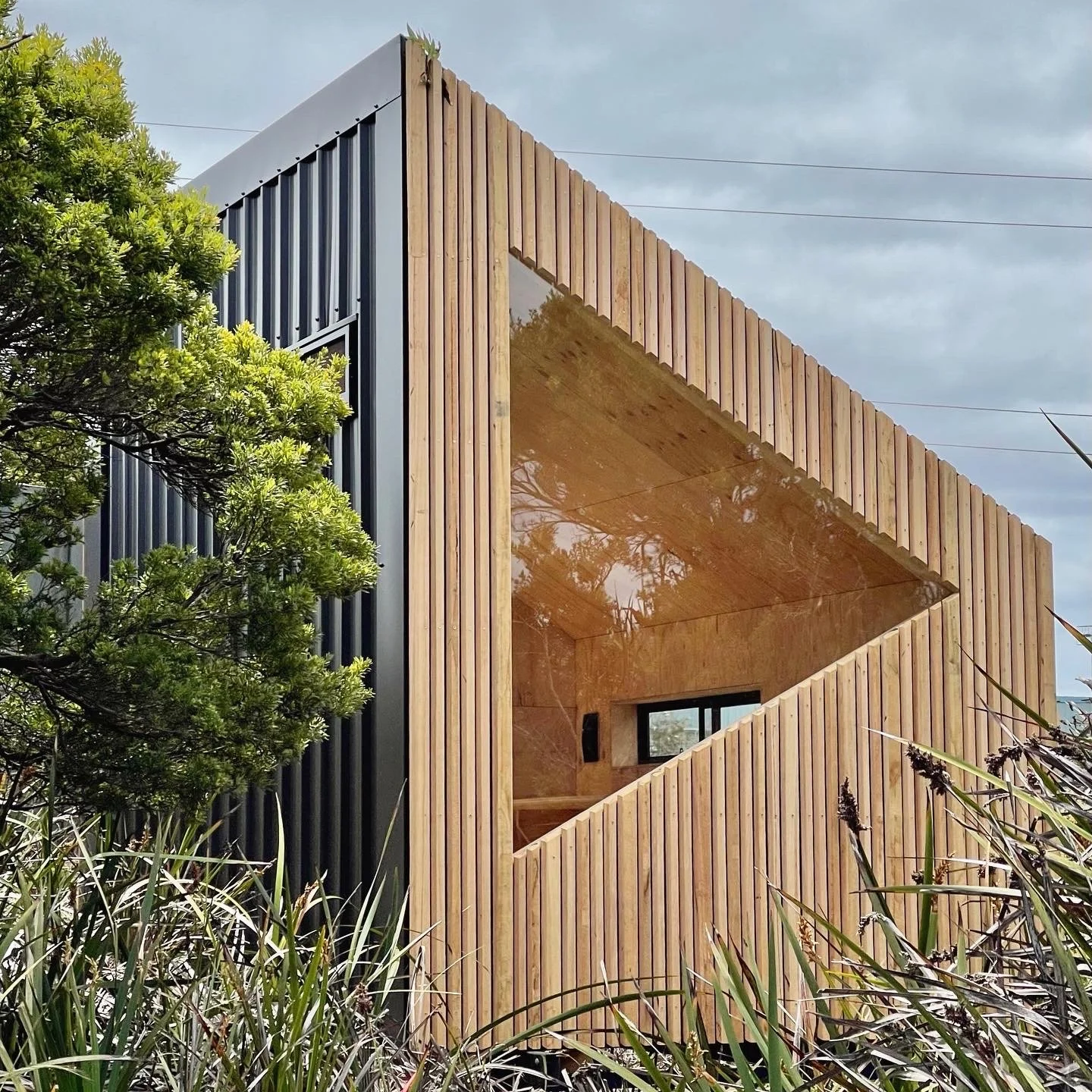 Modern architectural building with a unique angled wooden facade and large window, surrounded by greenery and tall grass, under a cloudy sky.