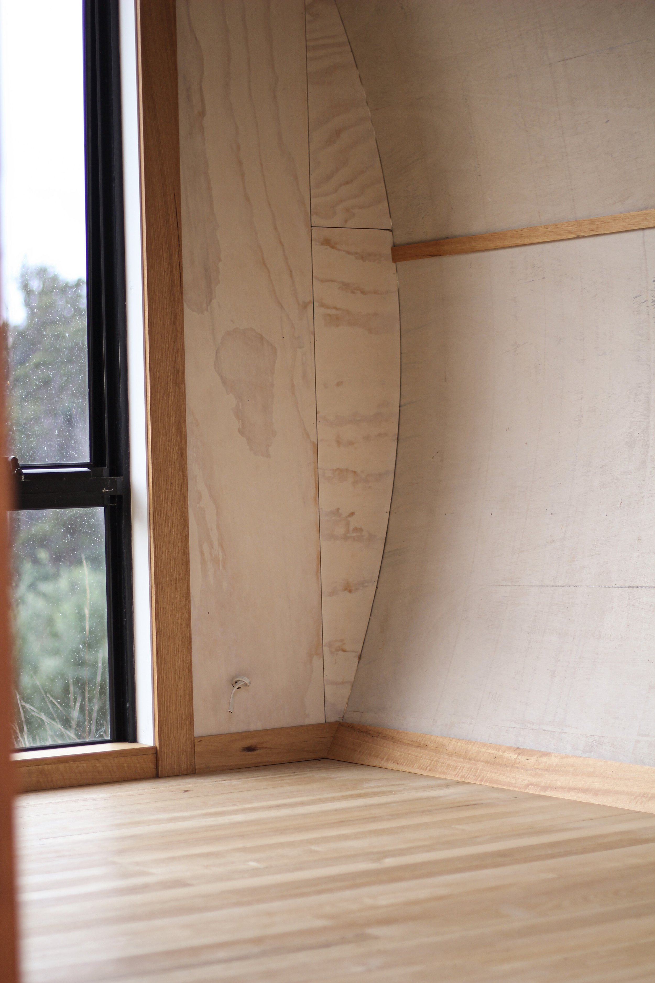 Interior corner of a room with wooden flooring, plywood walls, and a window showing an outdoor view of a cloudy sky and trees.