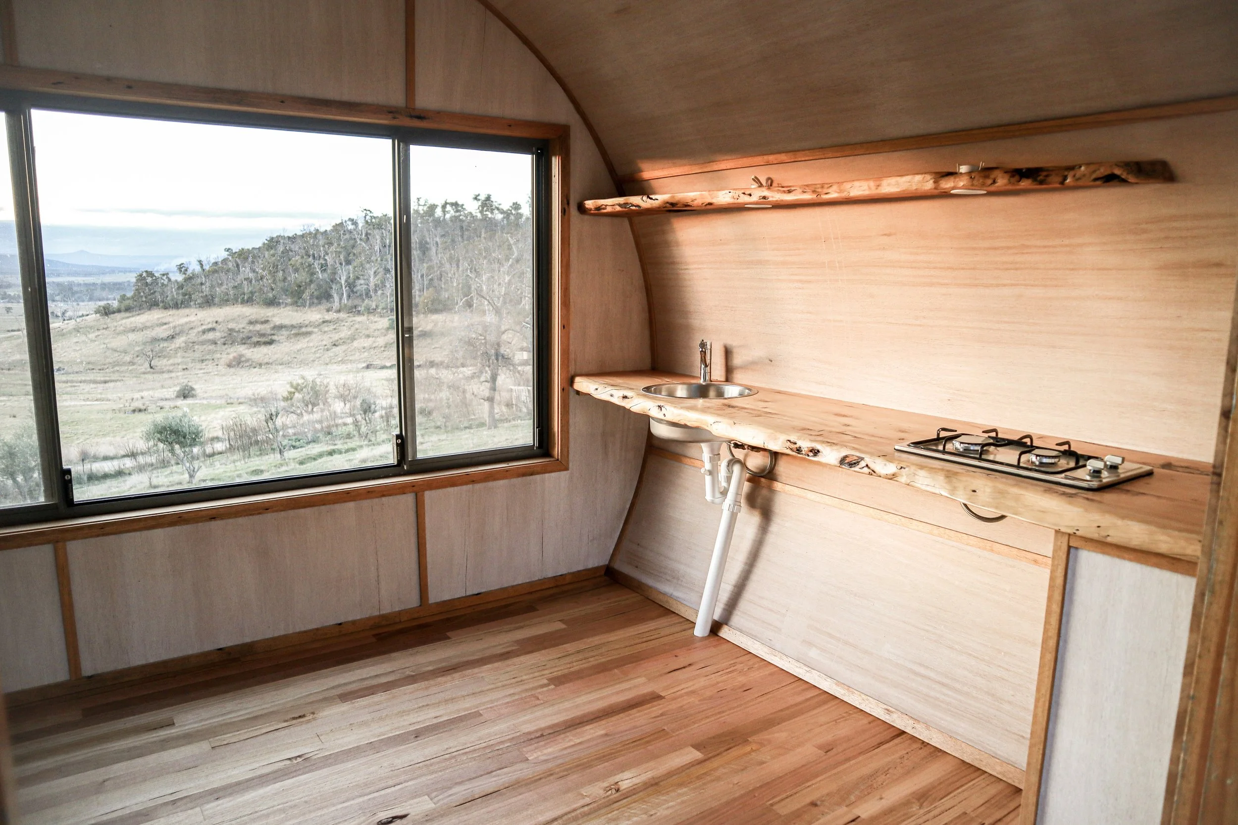 Small kitchen with wooden countertops and shelf, a small sink, a two-burner stove, and a large window overlooking a grassy landscape with trees.