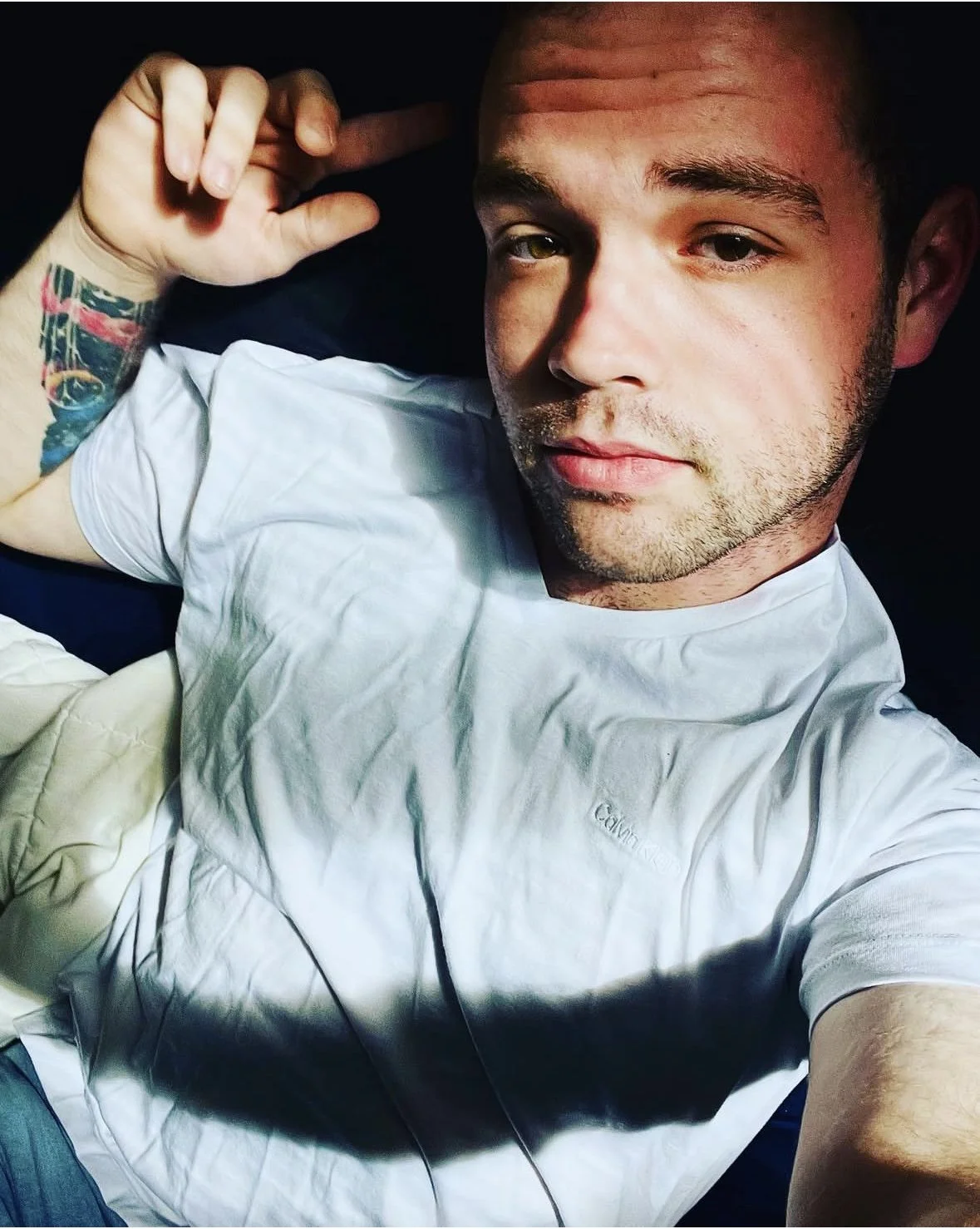 A close-up selfie of a young man with short hair, slight facial hair, and green eyes, lying down and wearing a white t-shirt. He is holding his hand near his head, with a colorful tattoo visible on his arm.