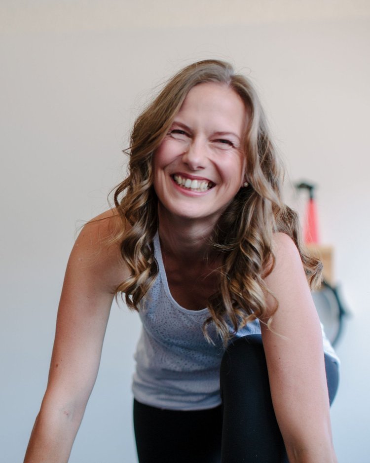 Maja Lund Loekkegaard Pilates Practitioner in pilates studio with long, wavy brown hair smiling and leaning forward in a casual setting.