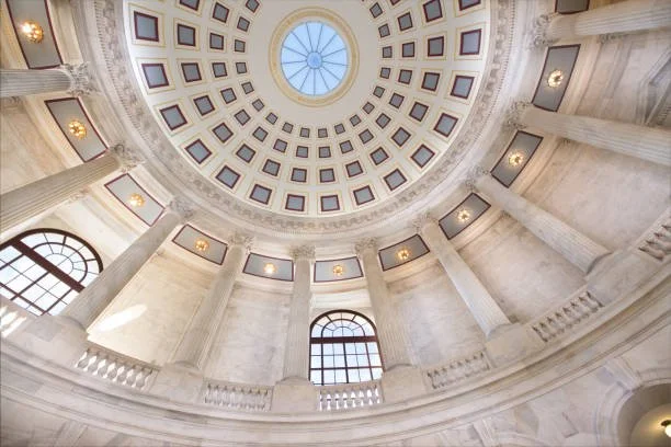 Interior view looking up at a domed ceiling with a central skylight, adorned with rectangular windows and classical columns in a grand building.