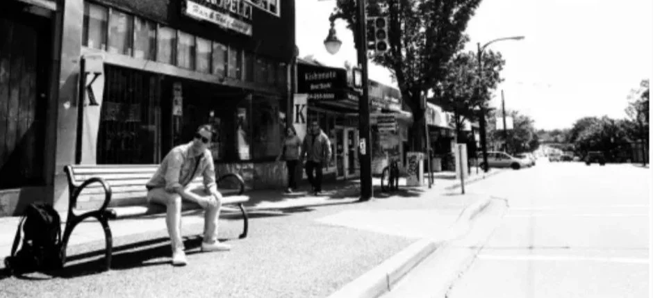 A black and white photo of a city sidewalk with a man sitting on a bench reading a phone, several people walking in the background, storefronts, and trees lining the sidewalk.