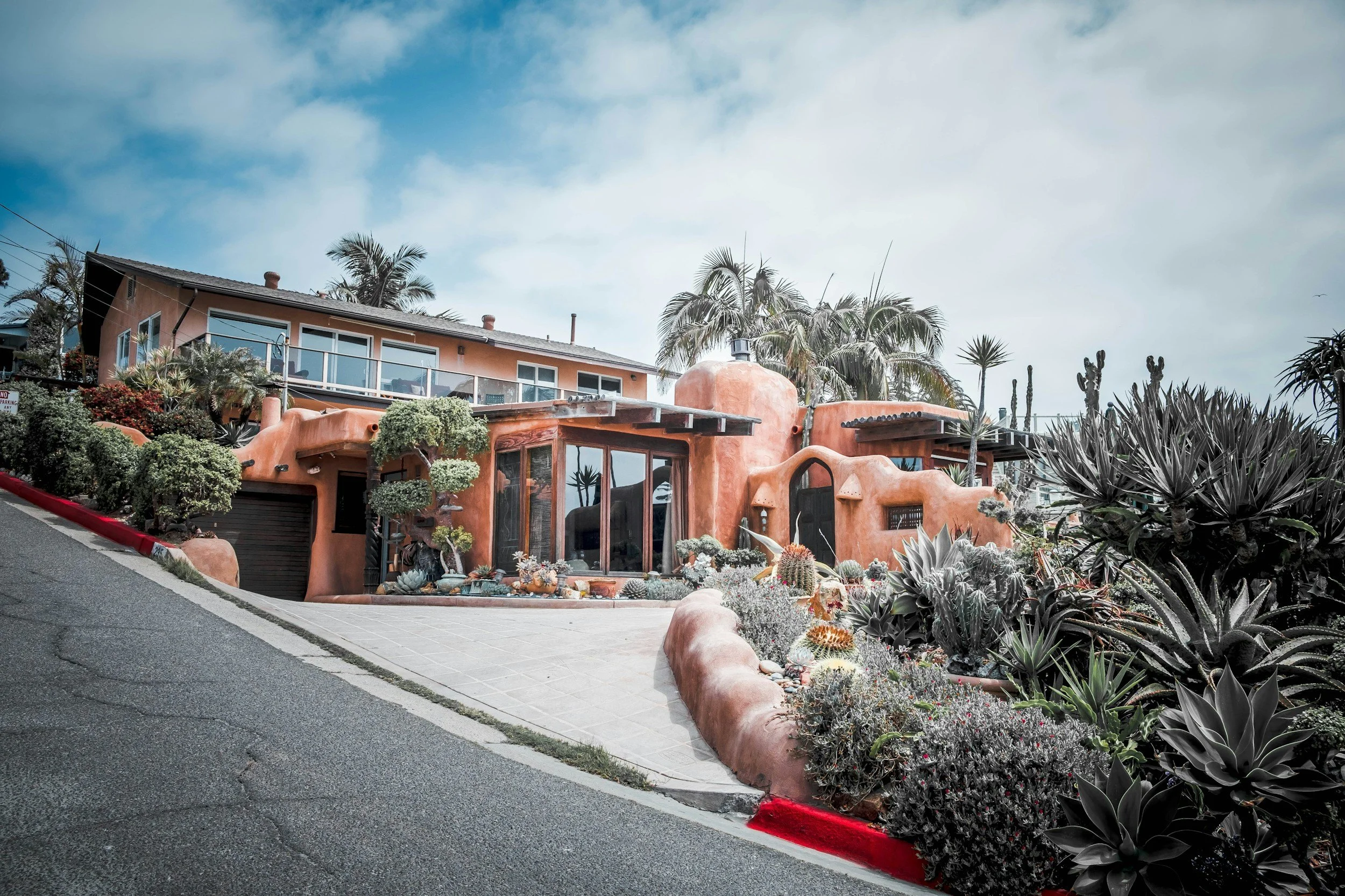 An Adobe-style house with rounded edges and a pinkish stucco exterior, surrounded by desert plants and cacti, on a hillside with a blue sky and some clouds overhead.