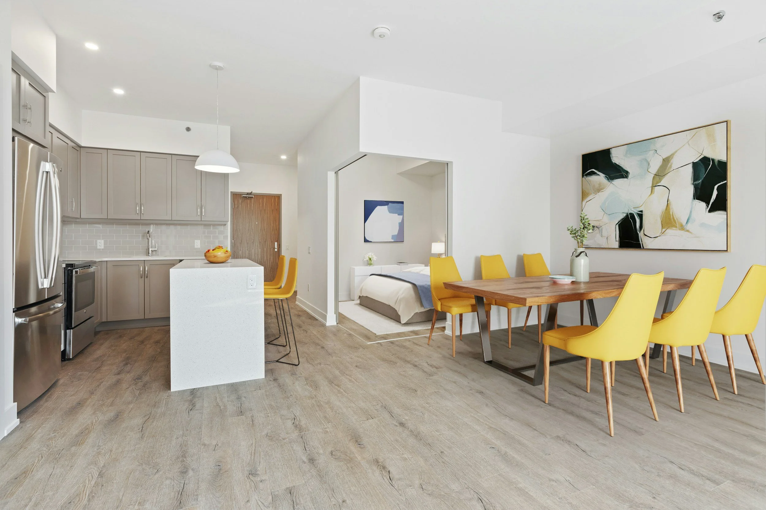 Open-concept kitchen and dining area with a view into a bedroom. The kitchen has gray cabinets, a white island with a fruit bowl, and stainless steel appliances. The dining table has a wooden top with yellow chairs, and there is a large abstract painting on the wall.