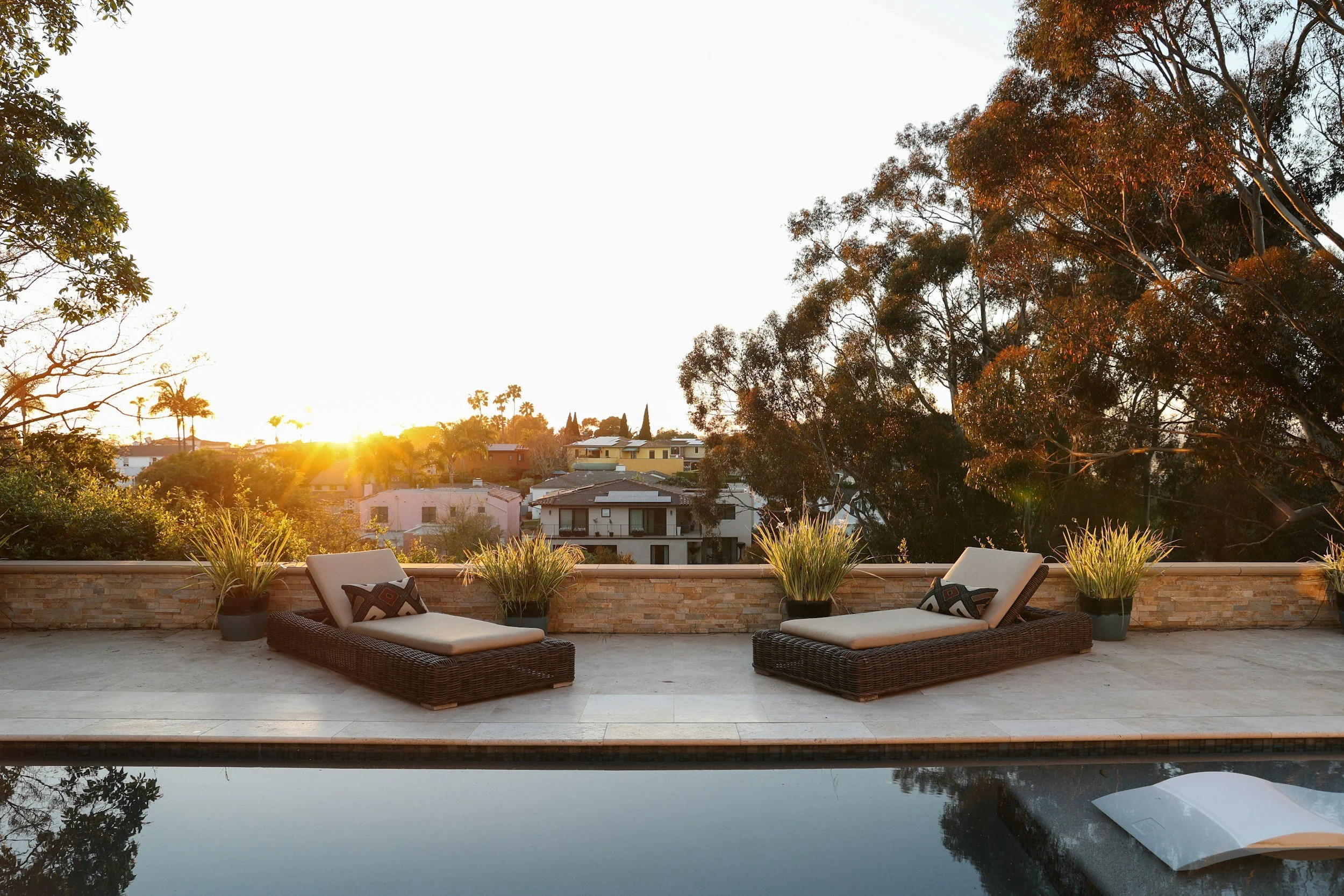 Outdoor patio with two lounge chairs, potted plants, and a view of a neighborhood with trees at sunset.