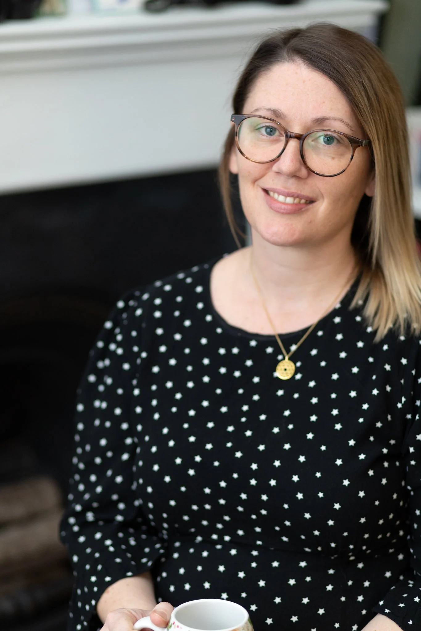 A woman with shoulder-length light brown hair, wearing glasses and a black dress with white star patterns, holds a white mug with a floral design. She is smiling and standing in front of a fireplace.