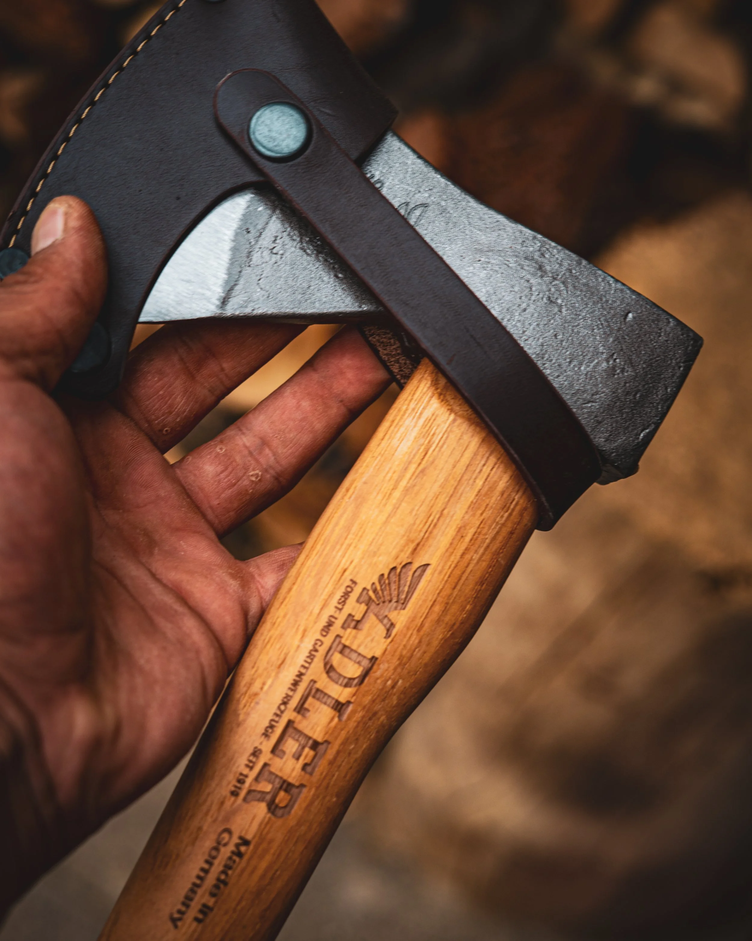 Close-up of a person's hand holding an axe with a wooden handle and a black metal head, outdoors with a blurred background.
