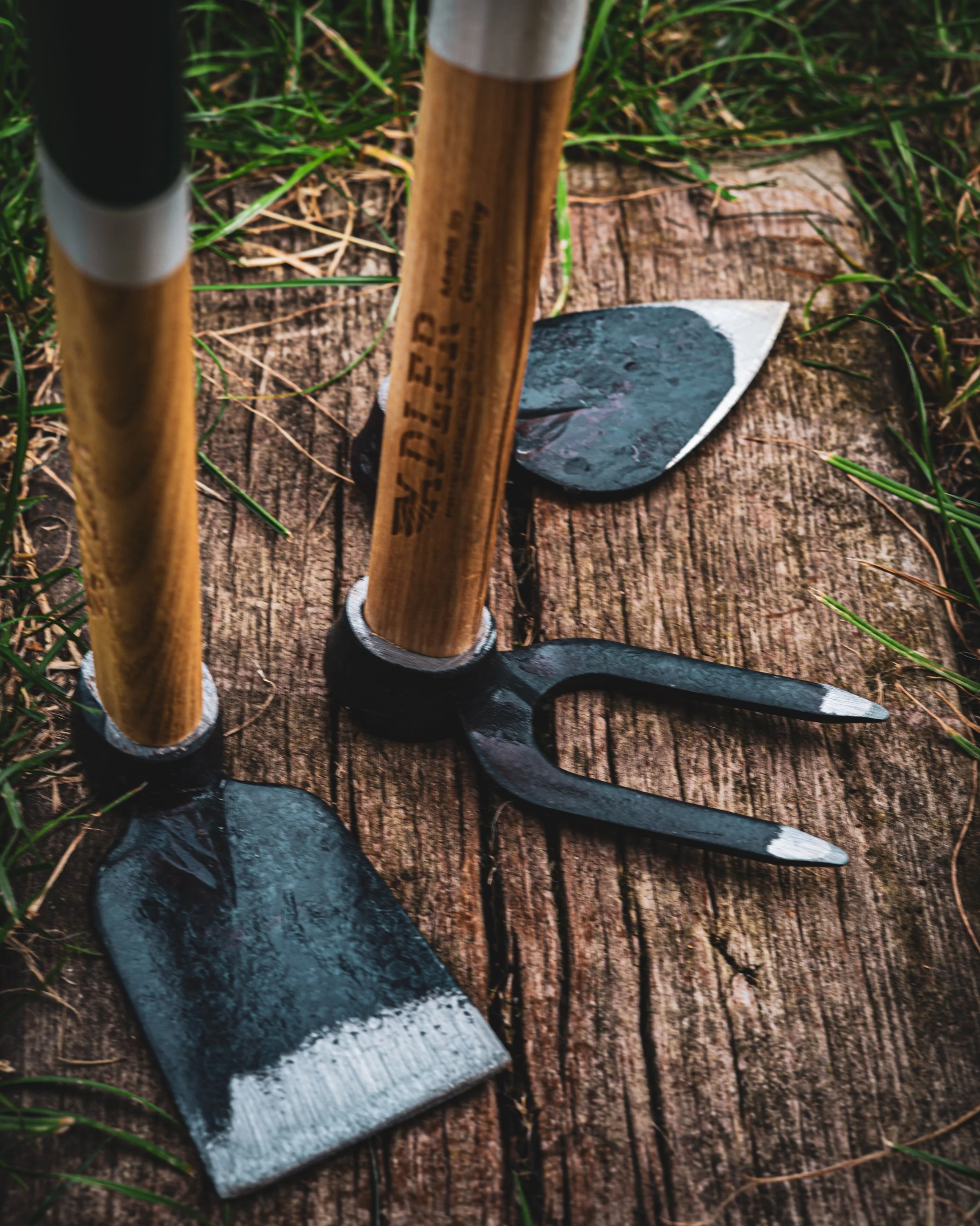 Close-up of gardening tools, including a small shovel, pruning shears, and a hand rake, placed on a weathered wooden surface outdoors surrounded by grass.