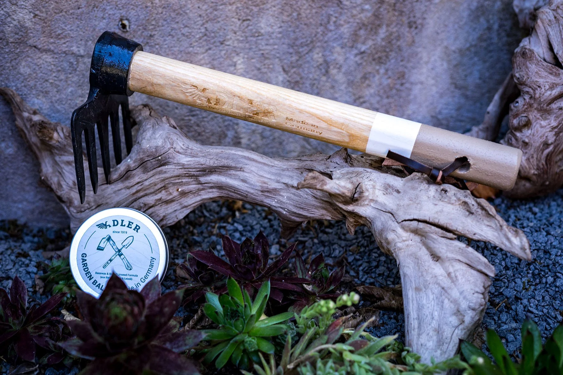 A gardening hand tool with a wooden handle and metal tines resting on a piece of driftwood among various green and reddish-leaved plants, with a container labeled 'ADLER' in the foreground.