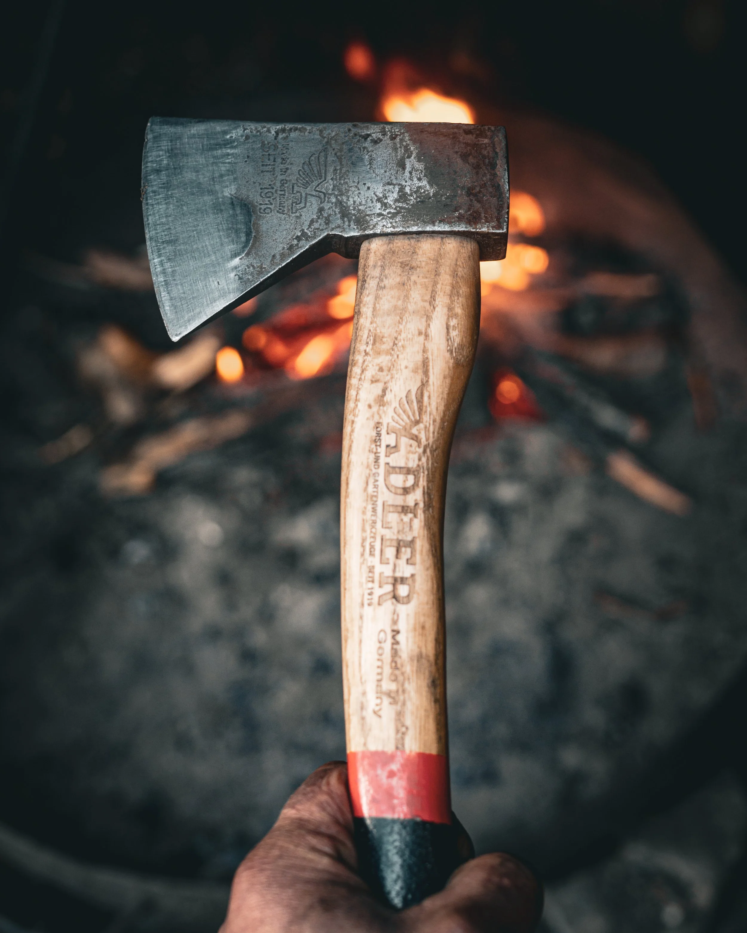 Close-up of an axe with a metal blade and wooden handle, held above a campfire with glowing embers and flames.