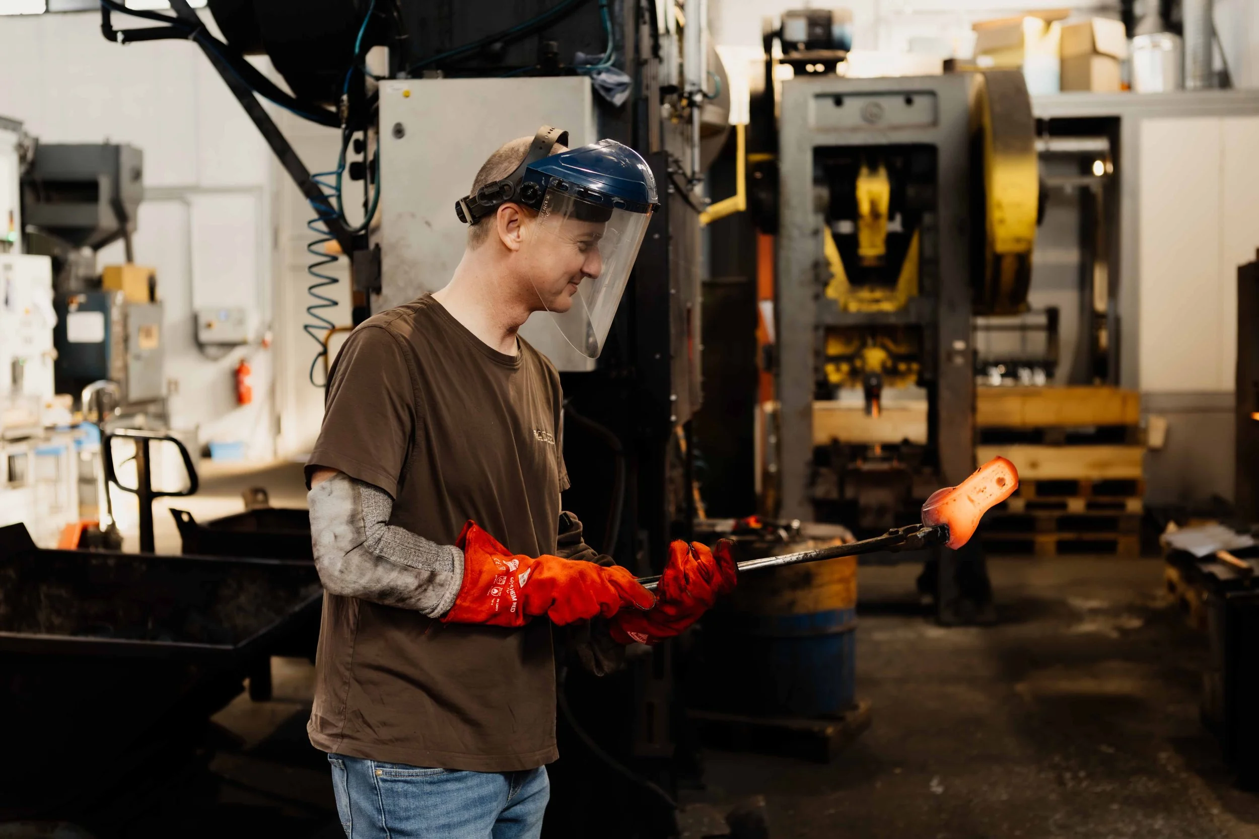 A worker wearing a face shield, gloves, and casual clothing is holding a heated metal rod with glowing orange molten material in an industrial workshop.