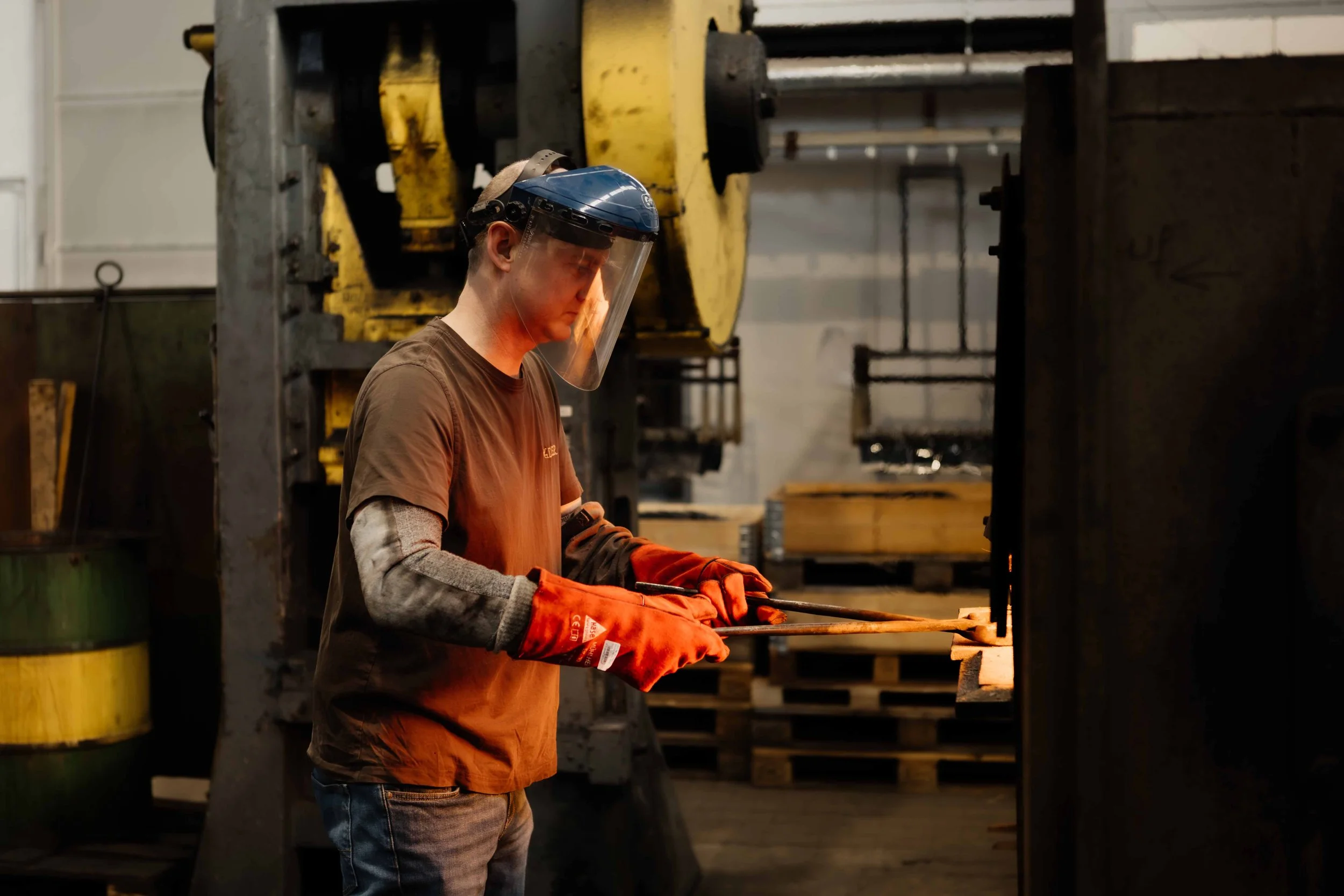 A worker wearing safety gloves, a face shield, and a brown shirt is operating a forging machine in an industrial workshop. The background includes large machinery and wooden pallets.