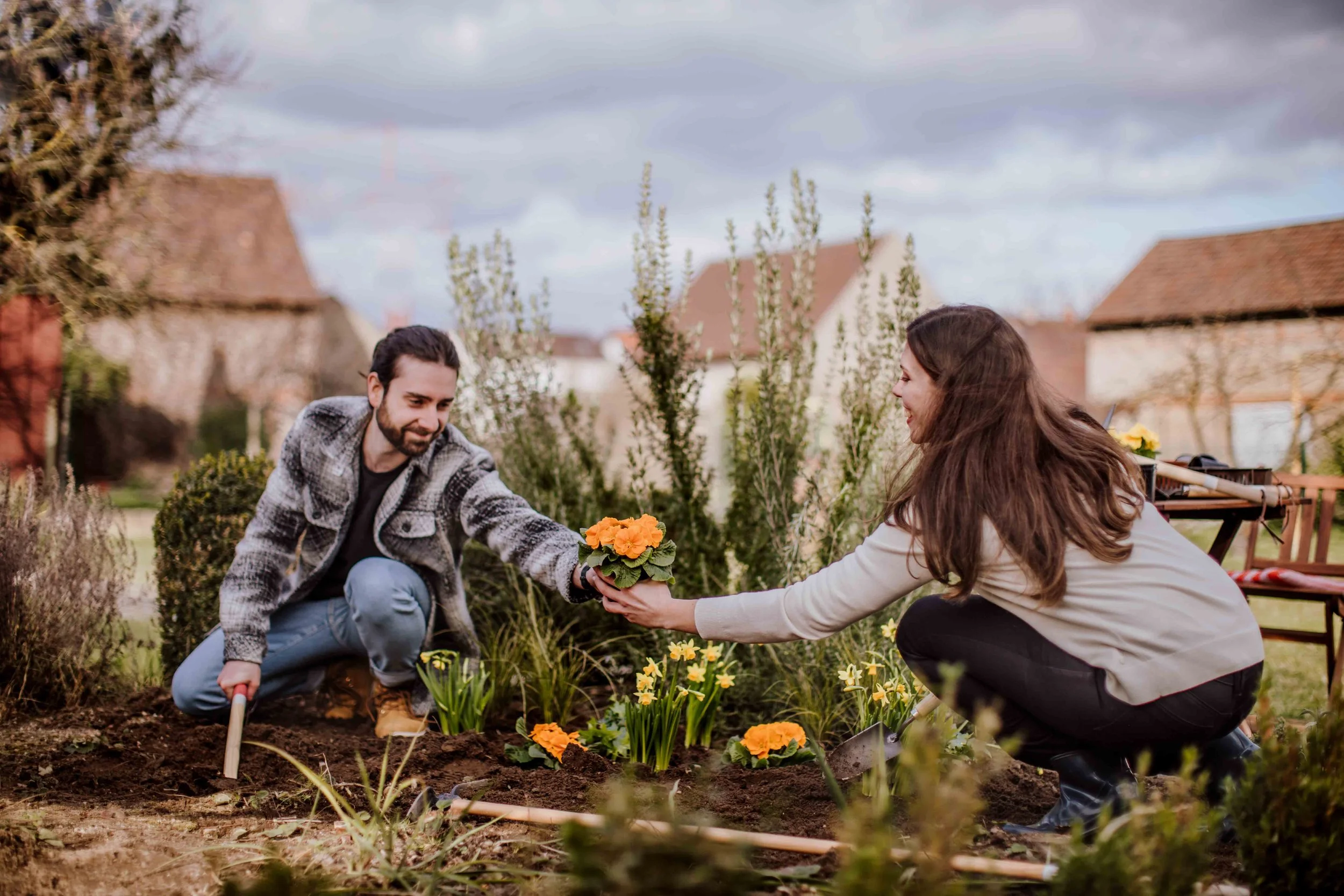 A man and woman gardening outdoors in a yard with flowers and by a shovelful of soil, with a background of houses and trees under a cloudy sky.