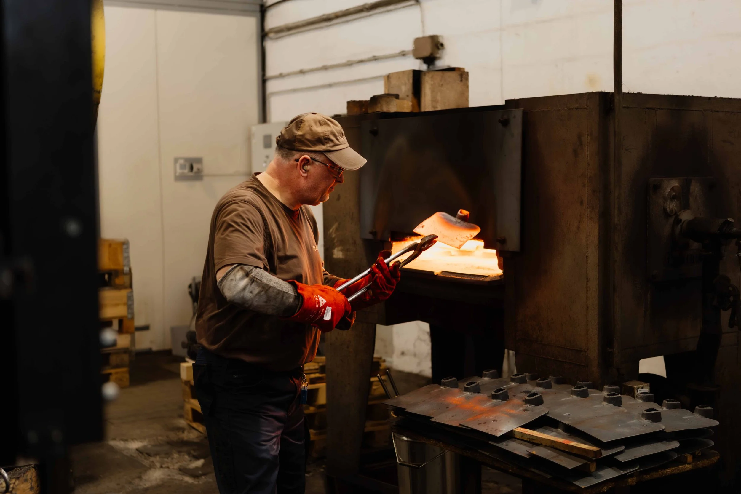 A blacksmith wearing safety glasses, gloves, and a cap, is holding tongs and working on glowing hot metal inside a forge.
