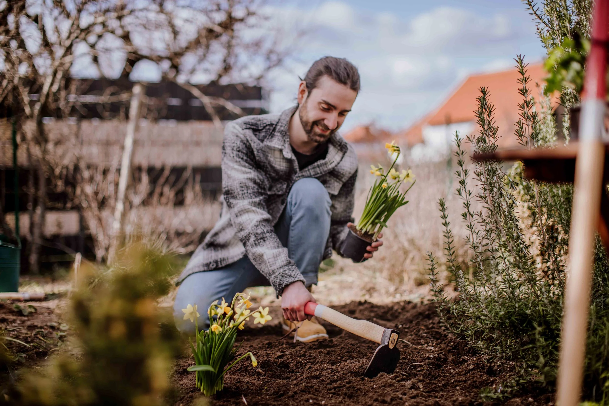 A man planting yellow flowers in a garden with a hand trowel, surrounded by plants and a wooden fence.