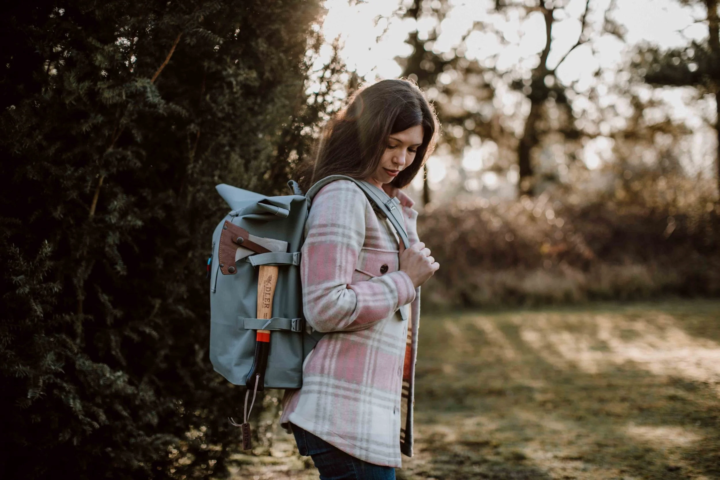 A woman with long dark hair, wearing a plaid coat, stands outside during sunset, with a large backpack on her back, looking down thoughtfully in a natural setting with trees and grass.