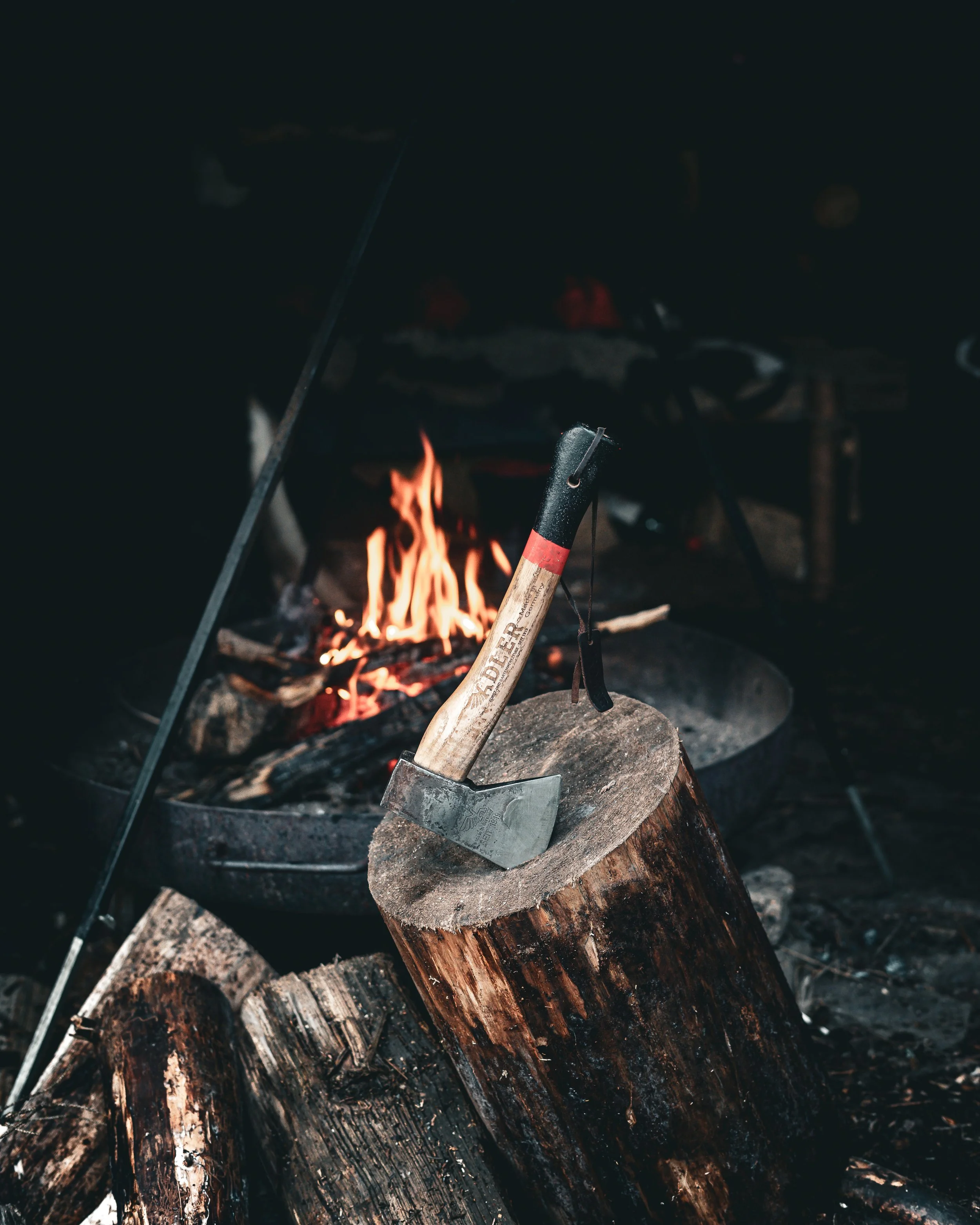 A axe embedded in a wood log in front of a campfire with flames, set outdoors.