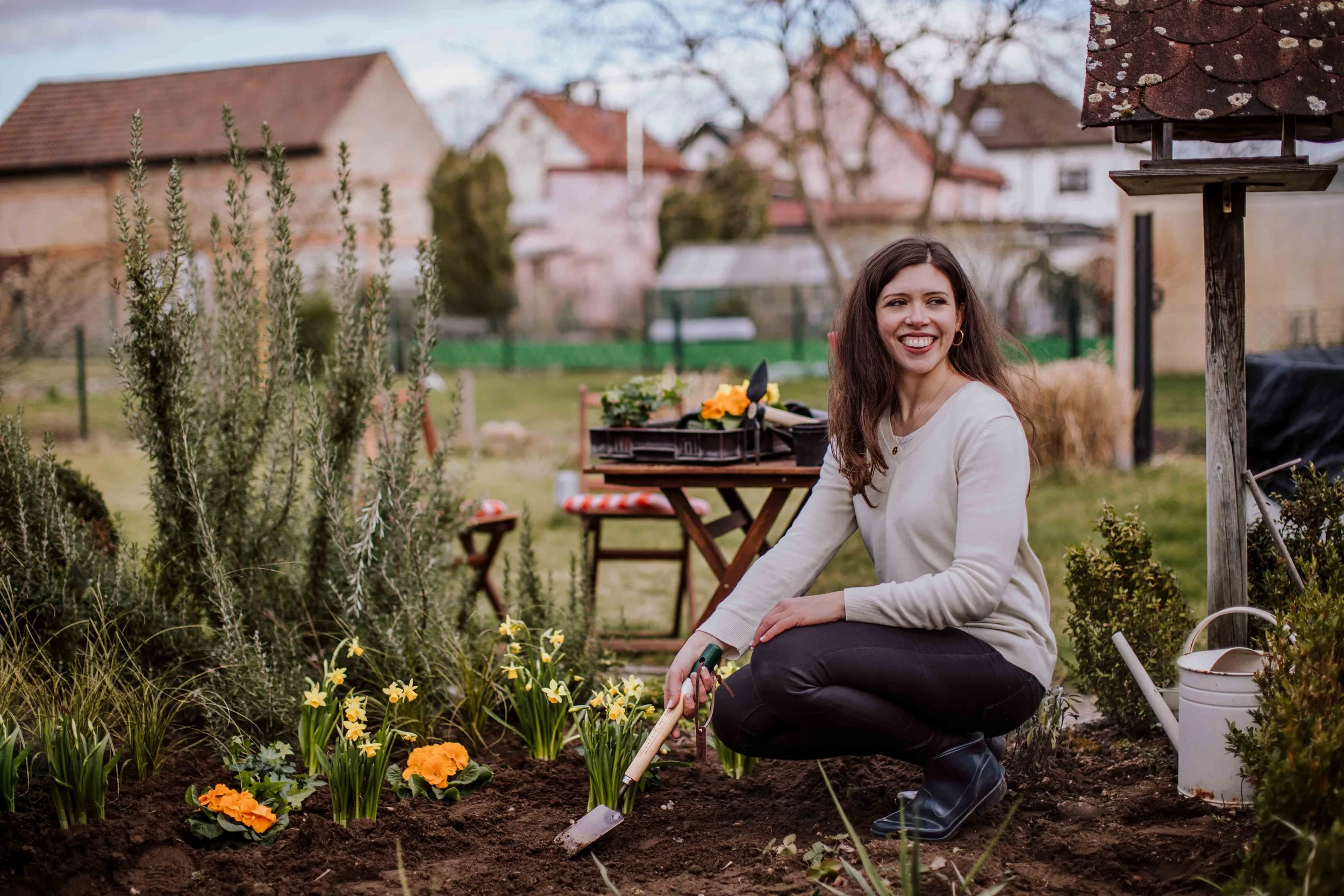 A woman planting flowers in a garden, surrounded by plants and gardening tools, smiling and squatting near freshly turned soil.