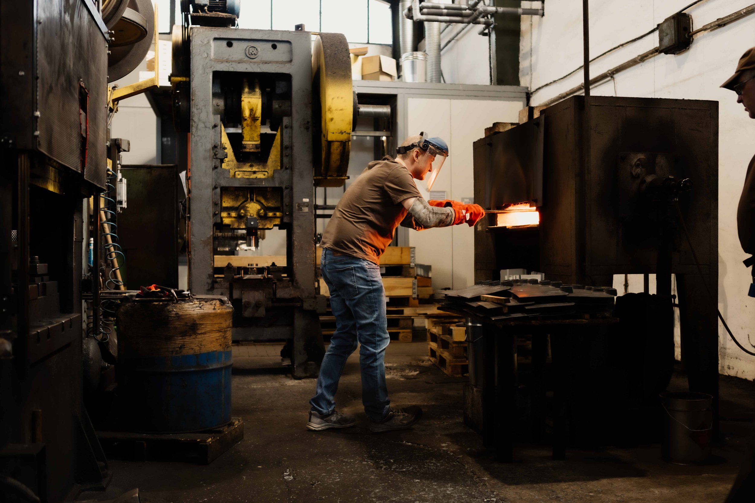 A worker in protective gear operating a forging press in an industrial workshop, with sparks and bright light coming from the heated metal.