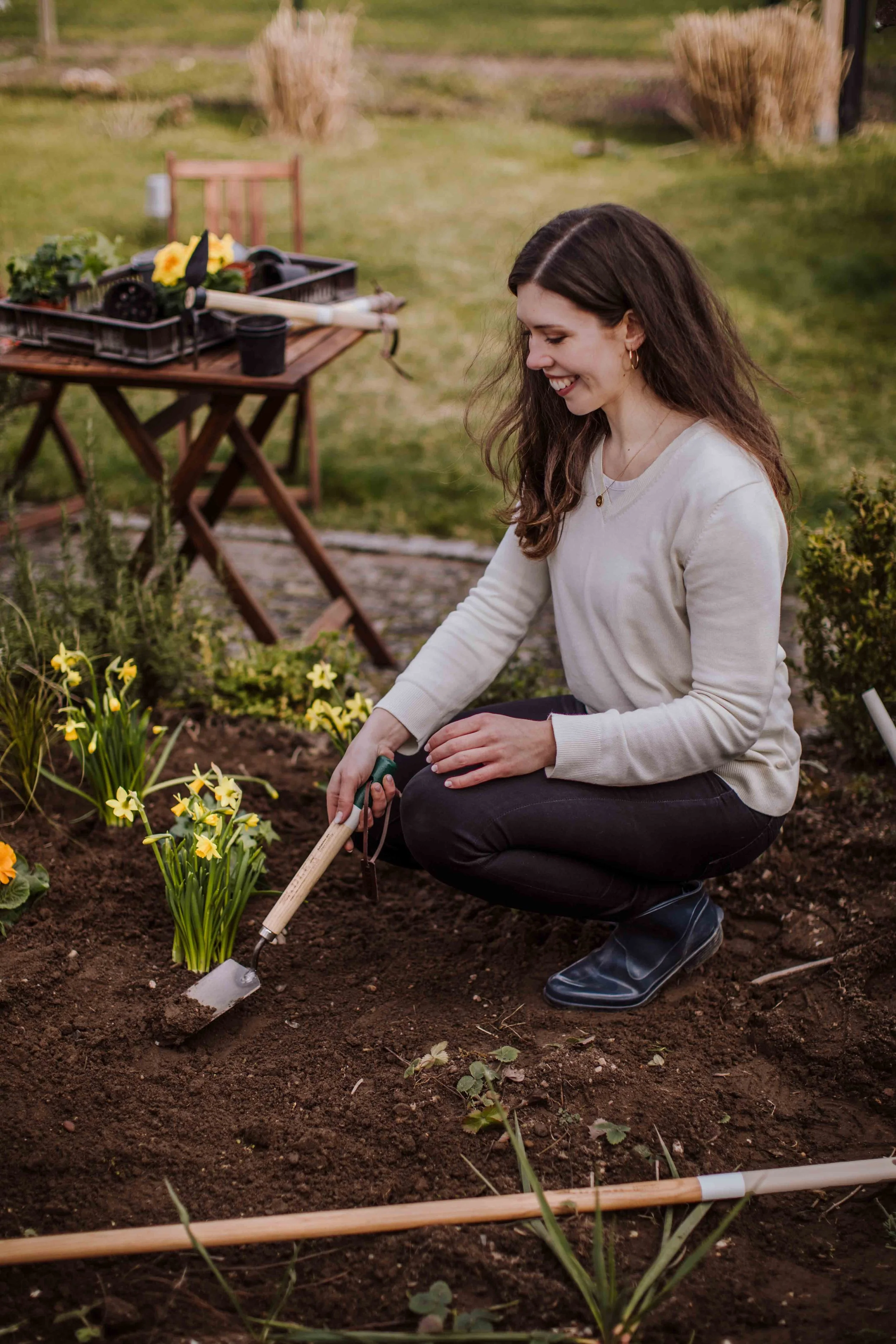 A woman planting yellow flowers in a garden, smiling while kneeling on the soil with gardening tools nearby.